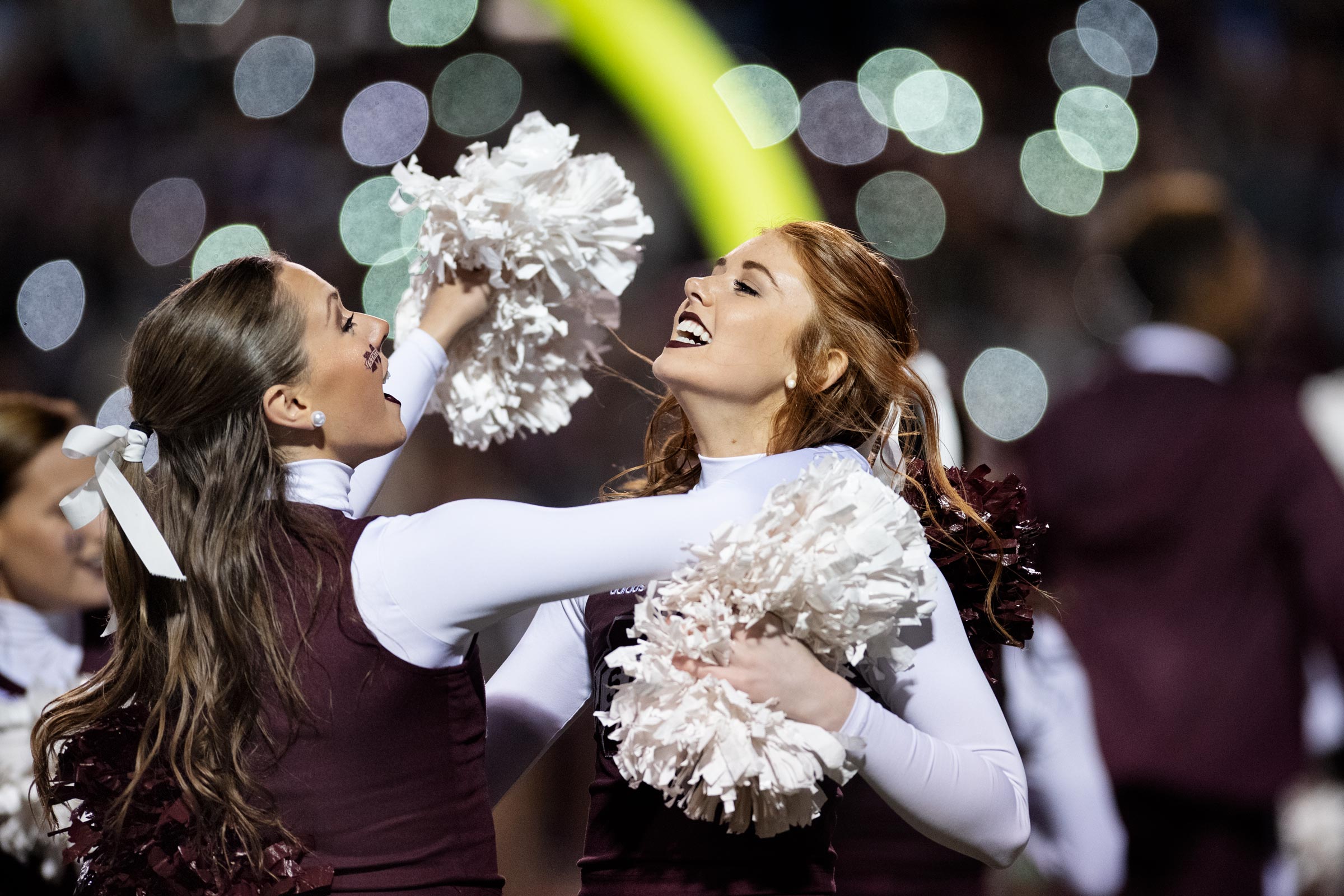 Cheearleaders hug and dance in celebration with sparkling lights of the stadium behind them.