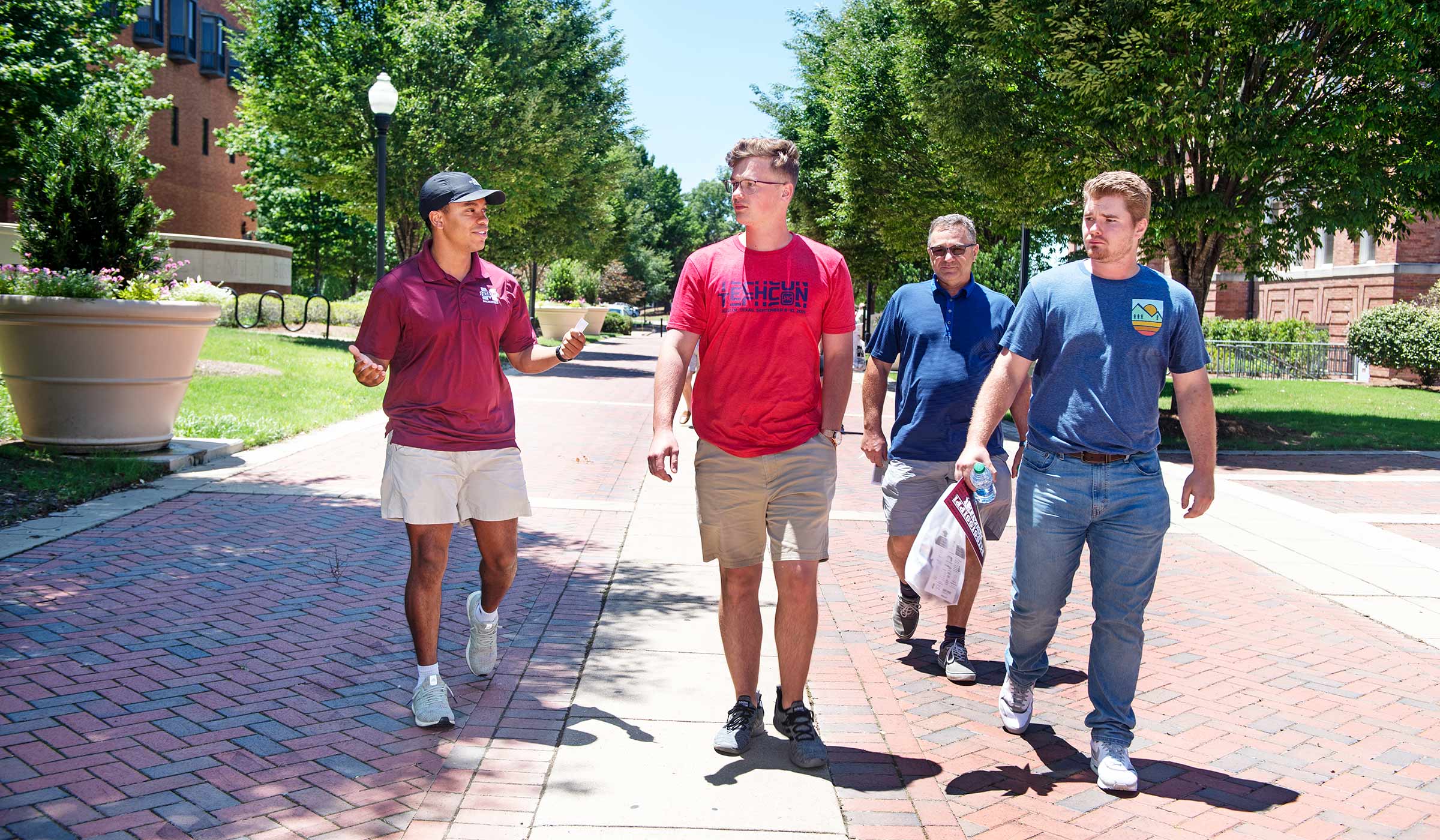 Student in baseball cap and maroon polo talking to three men on brick walkway