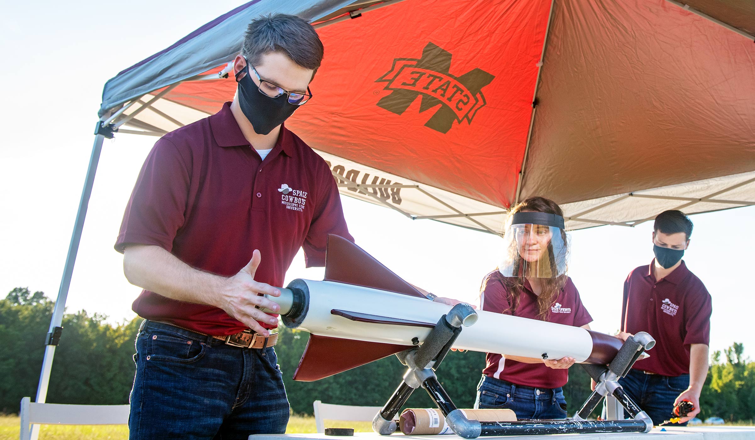 Two male students in maroon polos and masks and female student in face shield prepping rocket