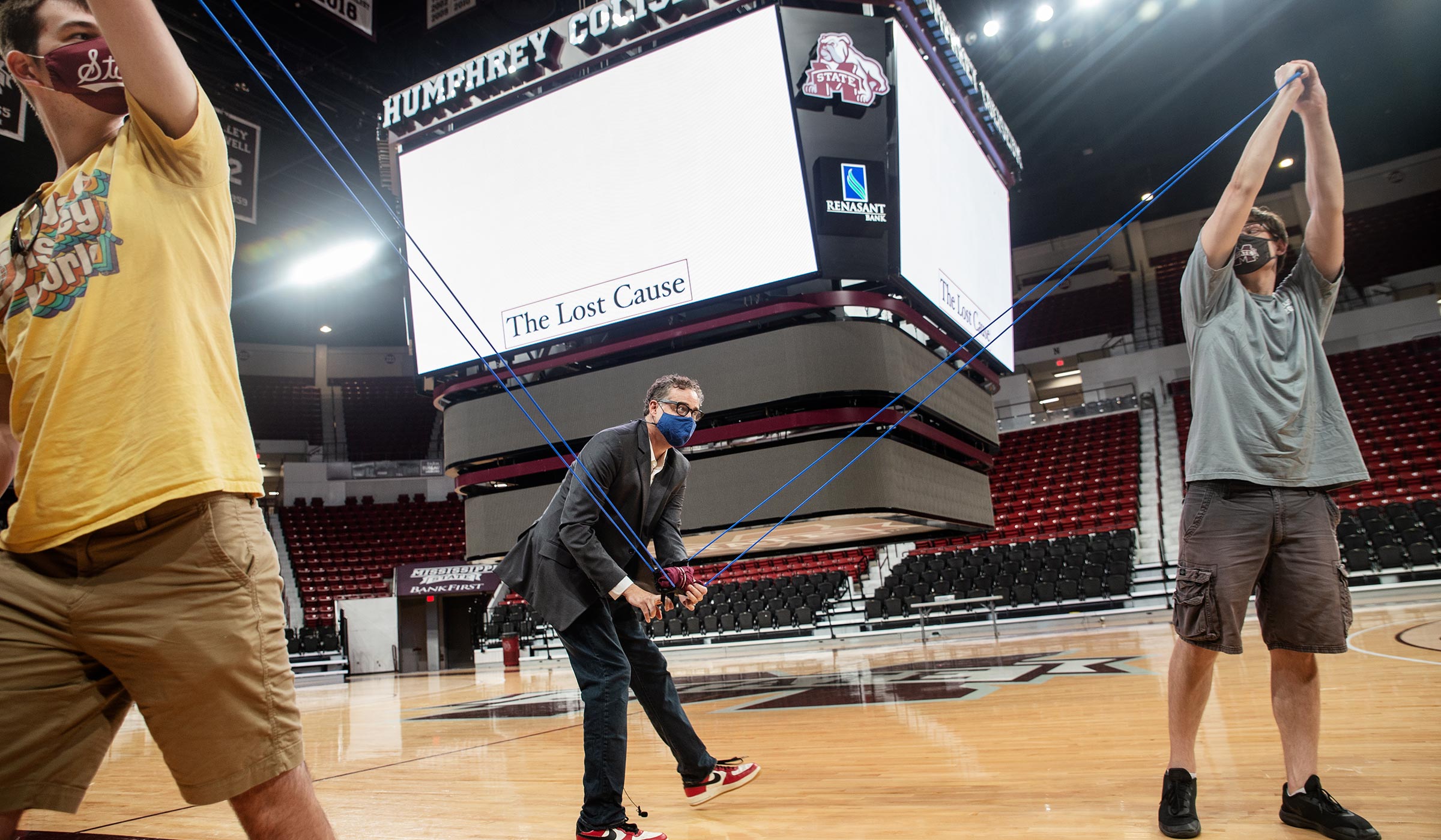 History Professor Jim Giesen pulls down the end of a giant slingshot in the Hump, framed by two masked students holding the ends.