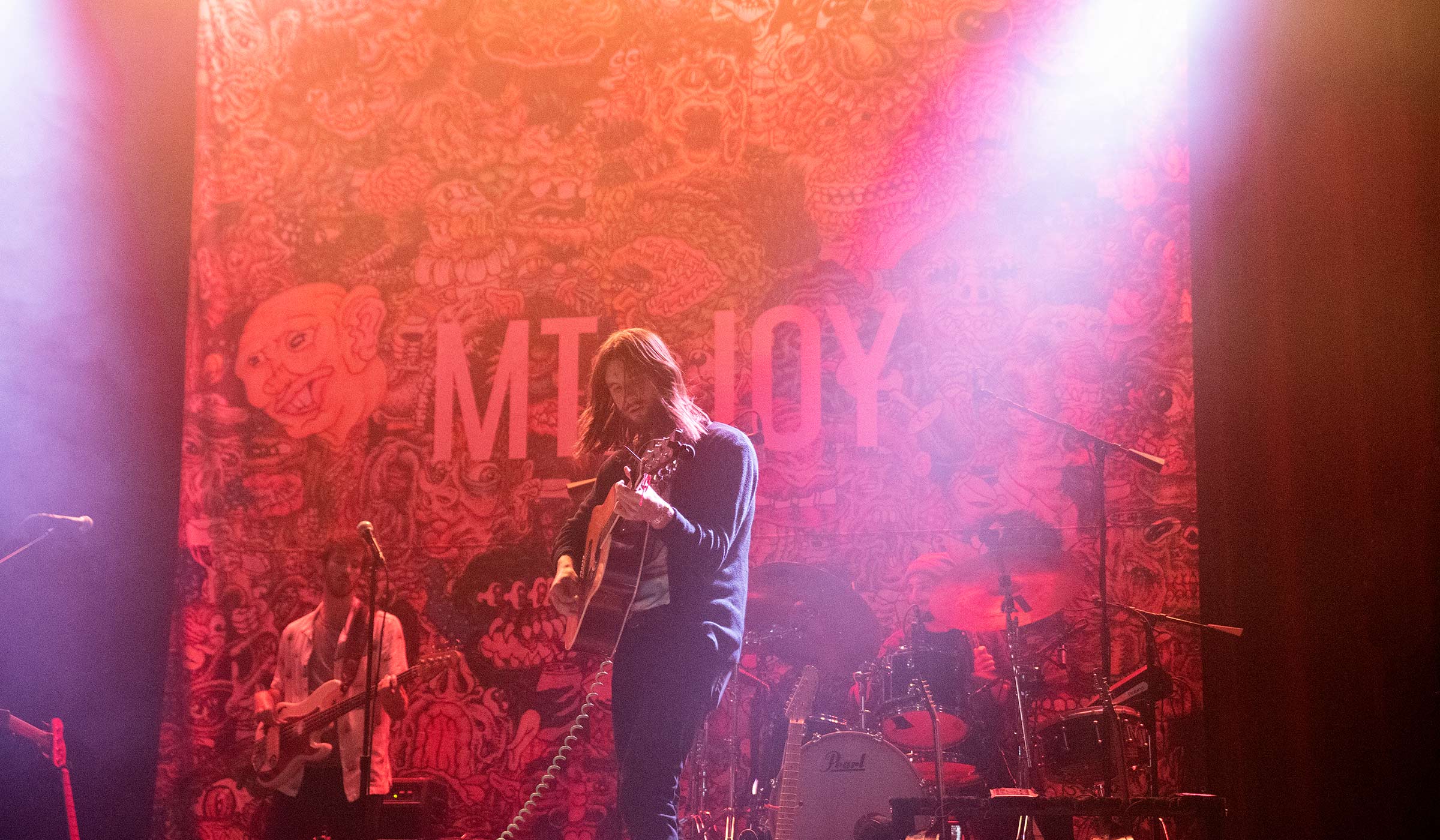 Man in foreground playing guitar on stage with pink and orange stage lights in front of artistic backdrop.