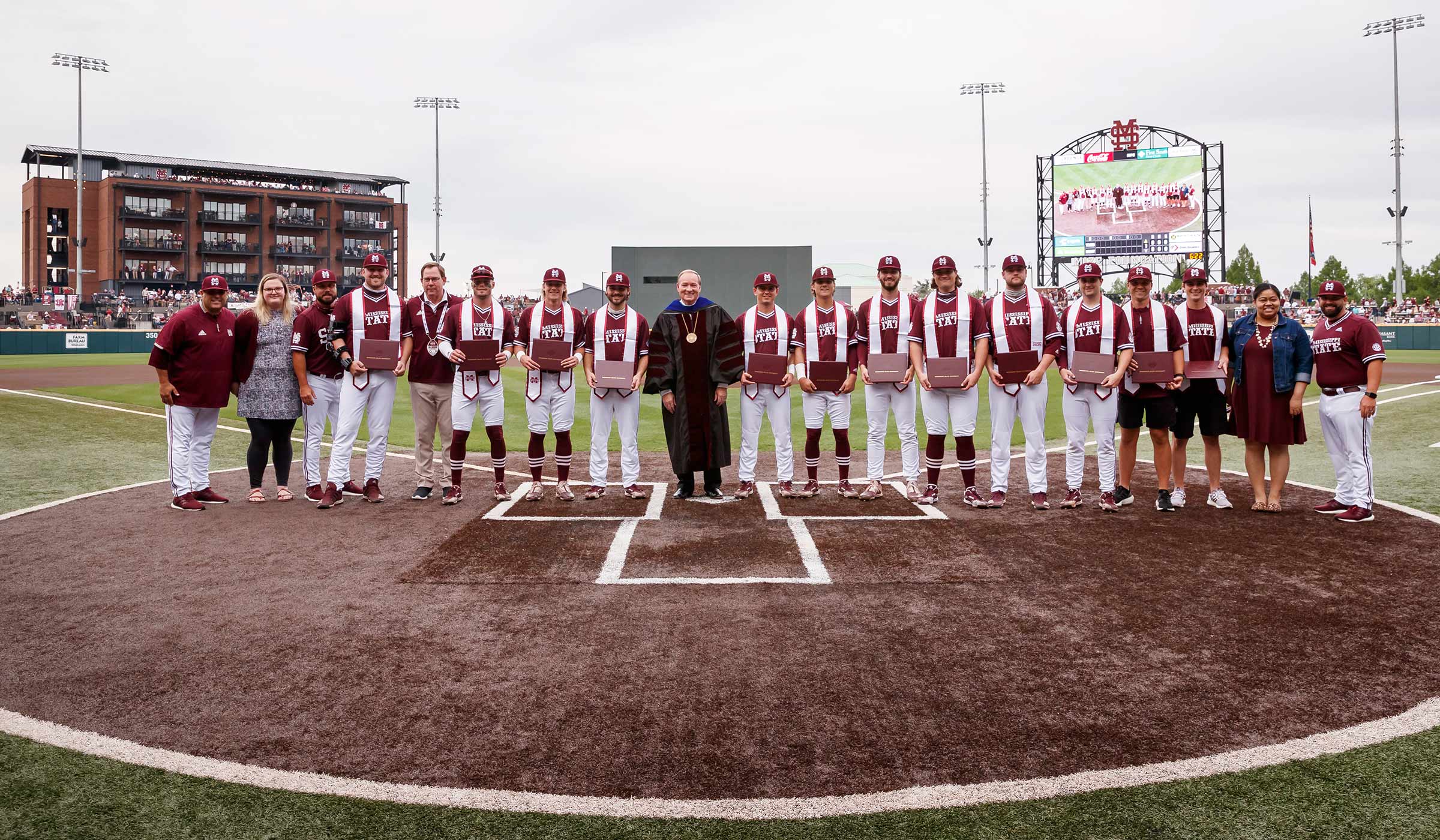 People lined up shoulder to shoulder some in maroon jerseys or tops with white pants holding diplomas