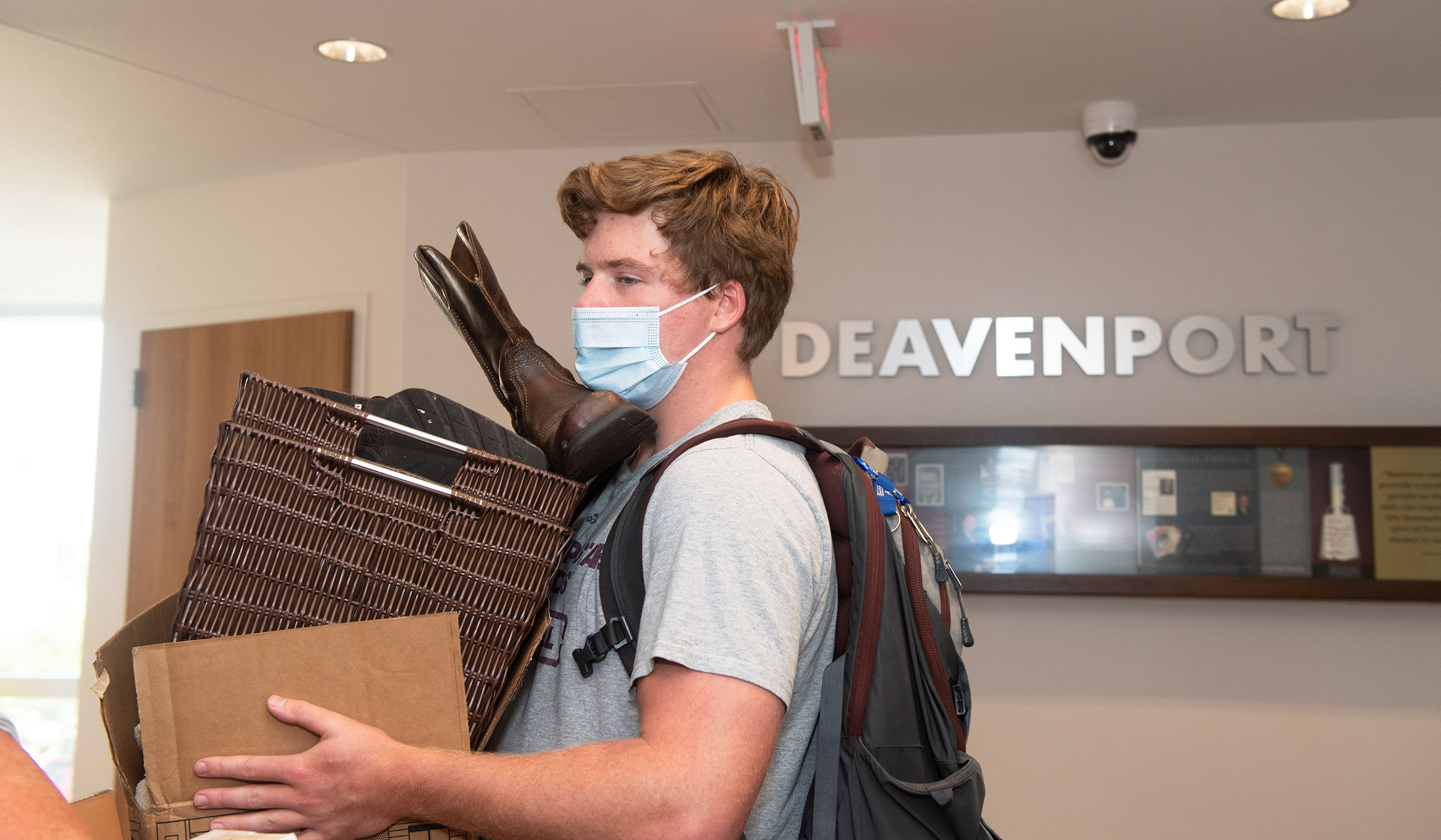 Male student in gray t-shirt and mask carrying boxes with boots