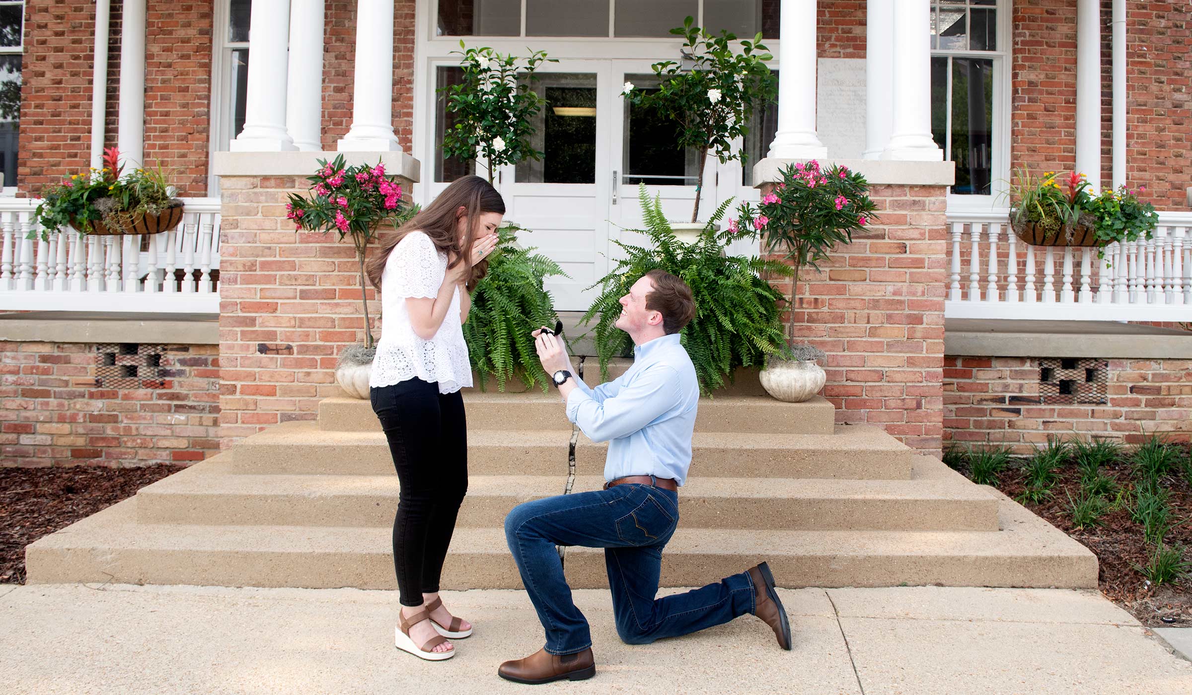 Man in jeans and blue shirt kneeling on one knee with ring box in front of female in white top and black pants