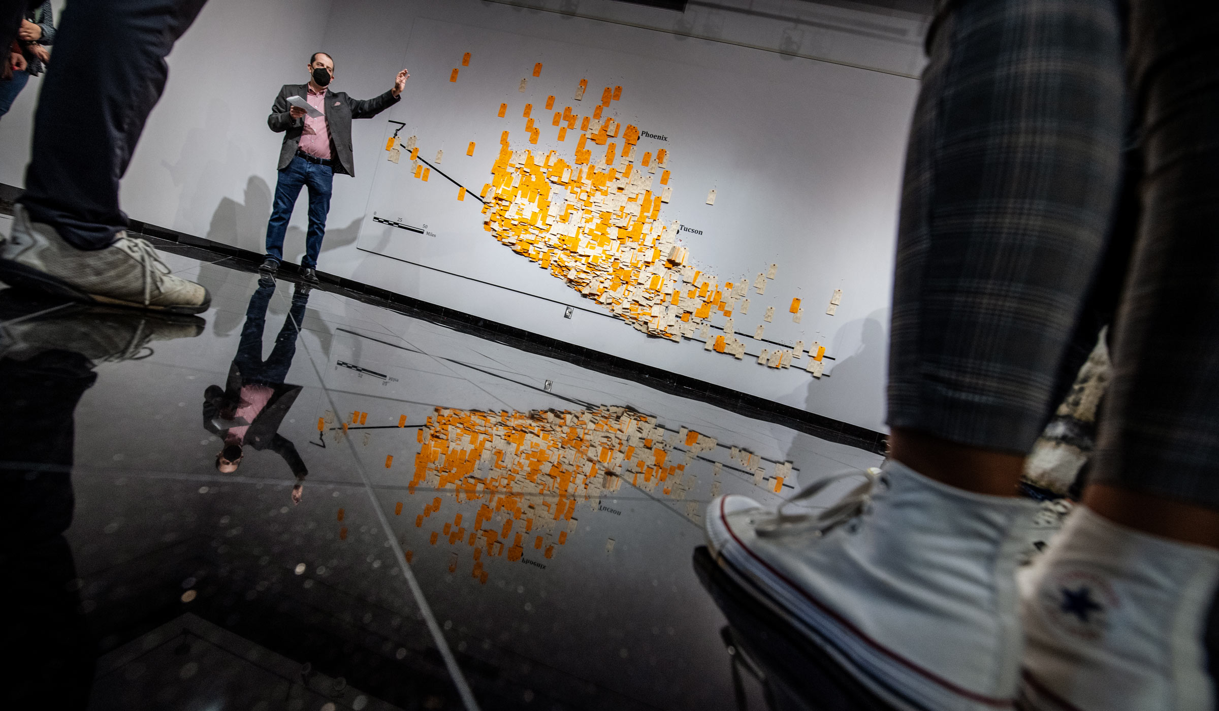 Framed by the feet of gallery opening attendees in the foreground, Anthropology professor David Hoffman speaks in front of the orange and manila Hostile Terrain 94 tags, which are pinned to the wall of the Colvard Union Gallery.