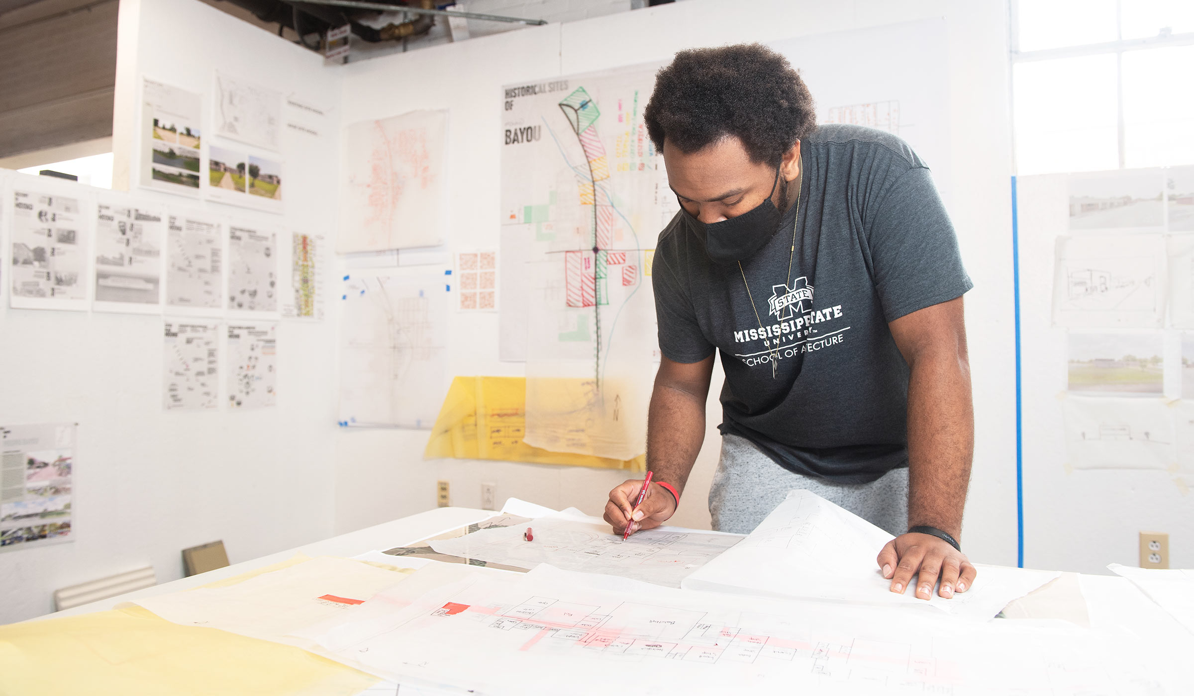 Male student in gray t-shirt and mask working on layout on his blueprint in front of wall covered in geographical plans