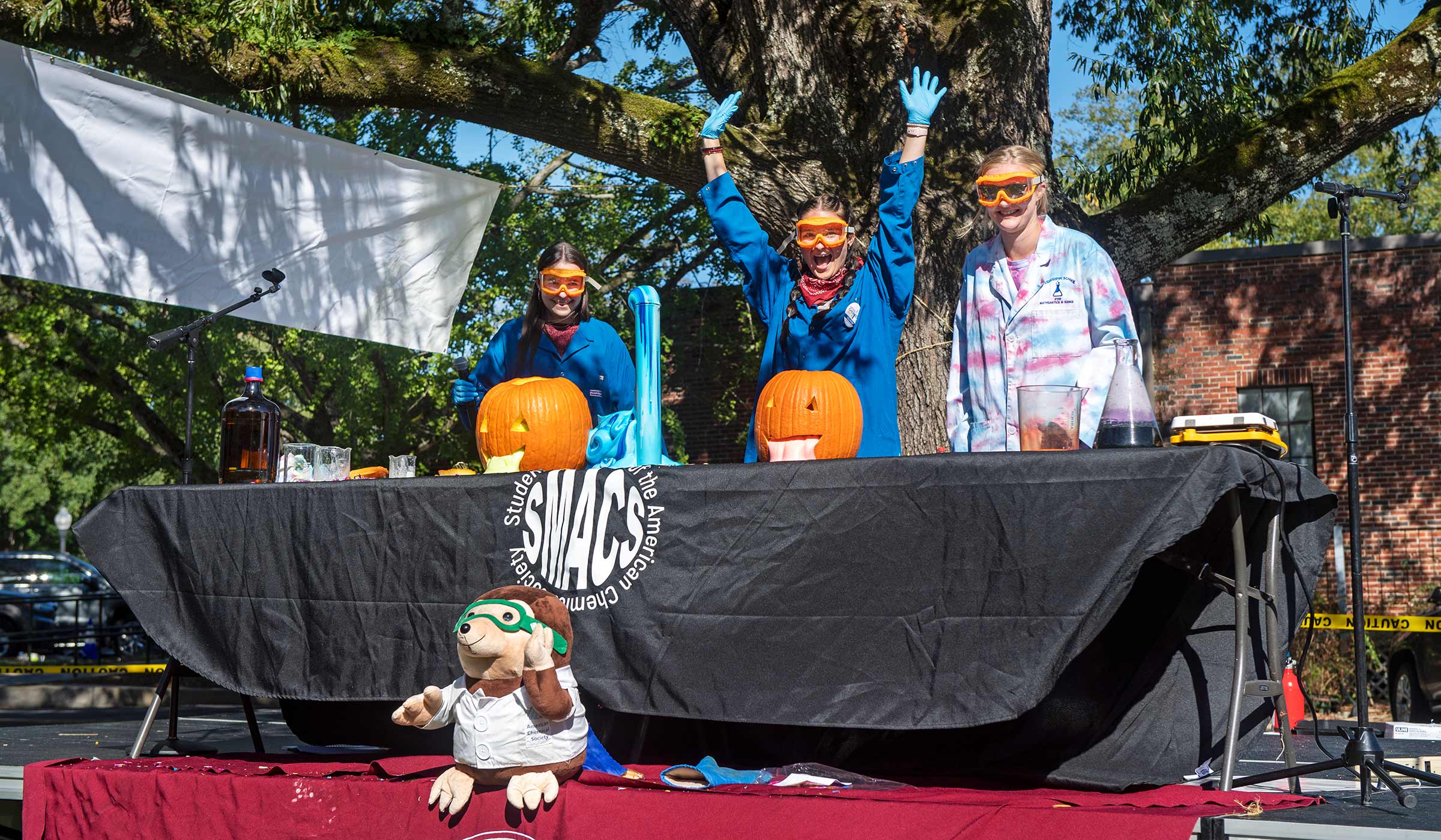 Female students in lab coats and goggles with pumpkins and large beaker oozing foam from chemistry experiment