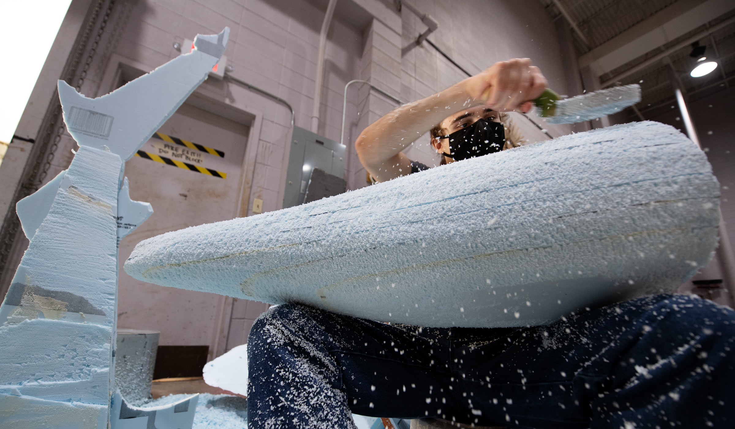 Student wearing mask uses scraping tool to shape blue form board into shape of dolphin tail.