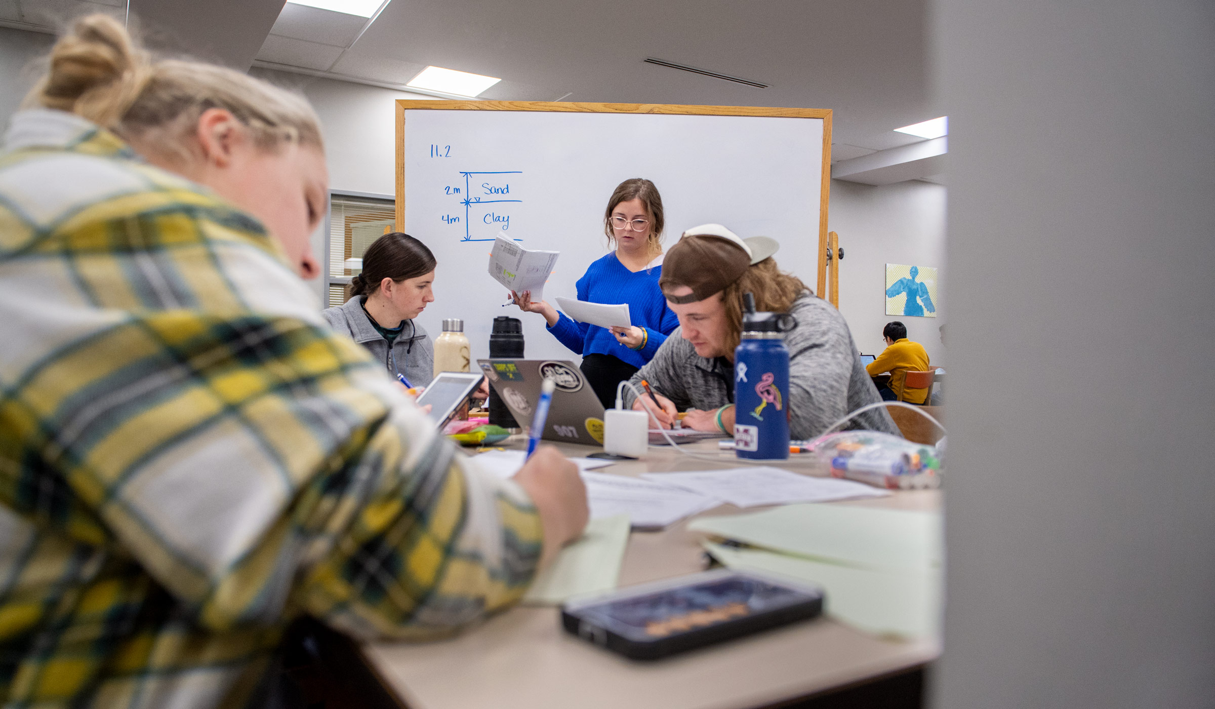 A group of students concentrates on their study materials around a table while one in blue readies to write on a whiteboad beyond.