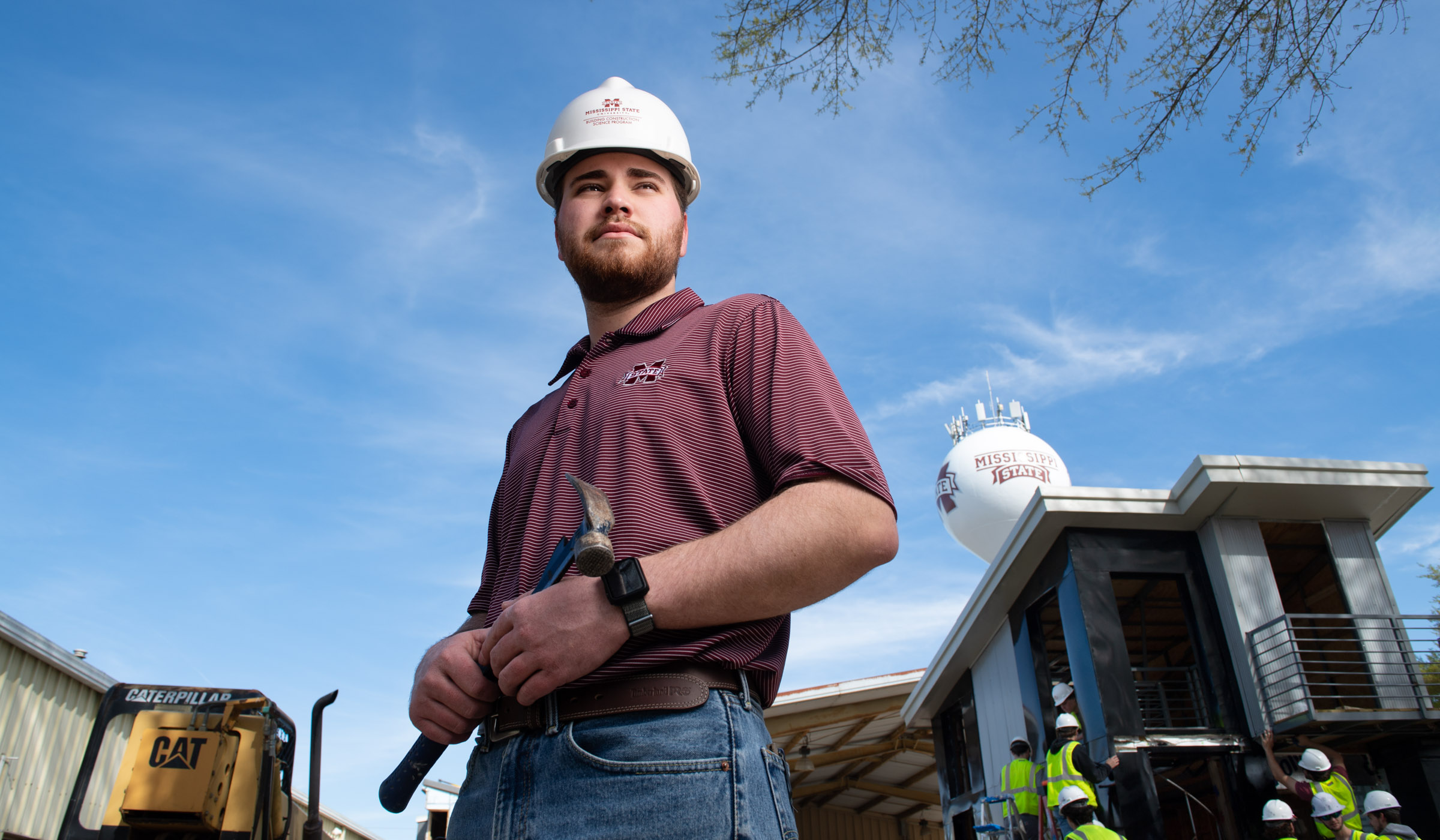 Jack Blacklock stands outside wearing an MSU shirt and hard hat, with construction of a tiny house in the background