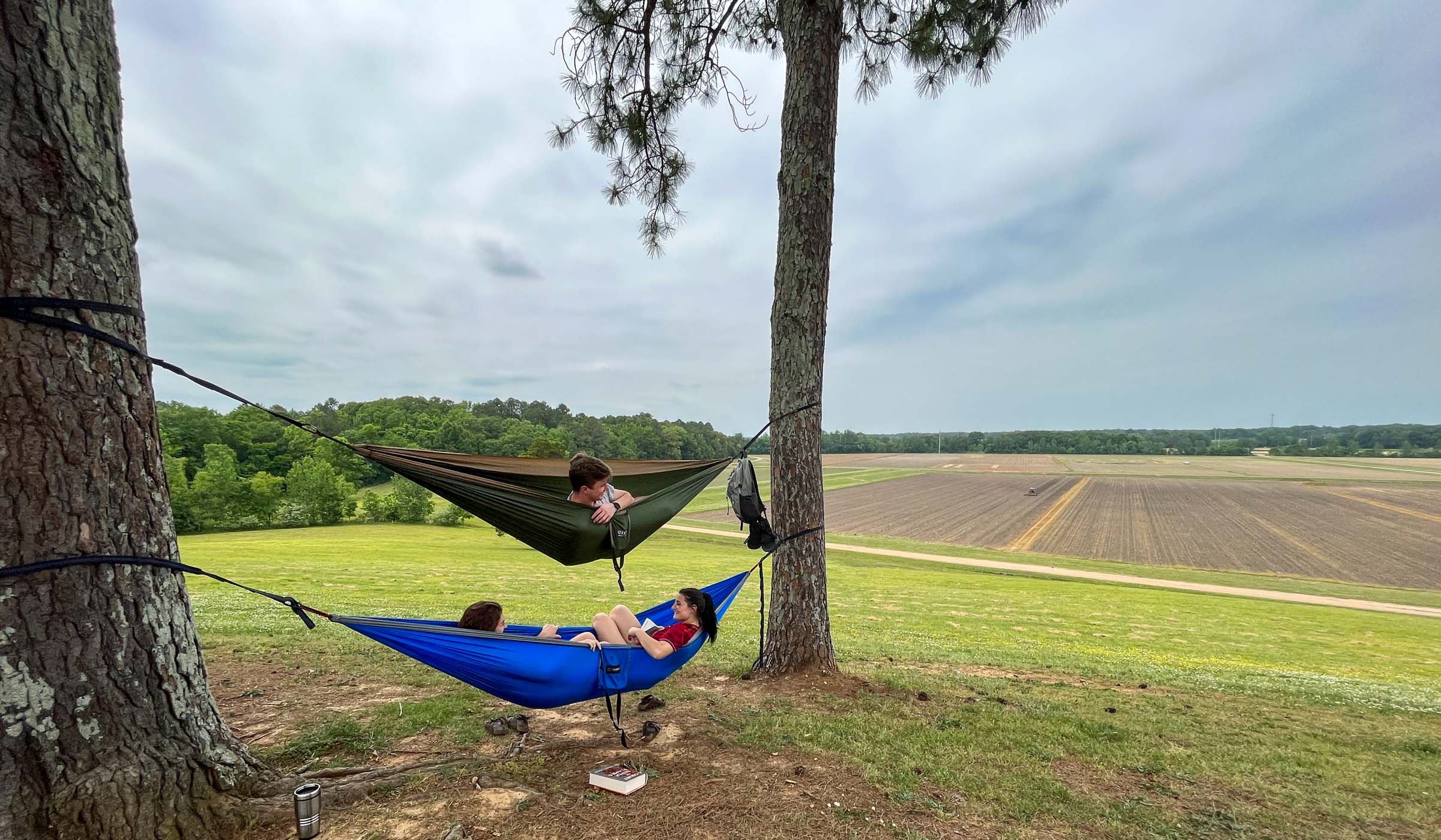 Three students relax in two hammocks hung between hilltop trees with views down over freshly plowed North Farm fields.