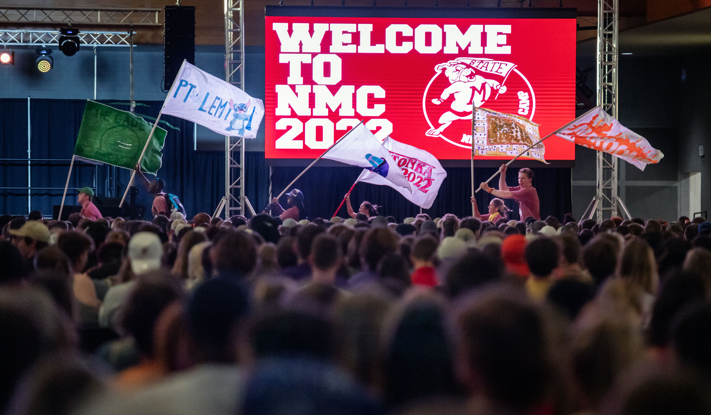 With a sea of New Maroon Camp attendees heads in the foreground, NMC staff run past giant screens with the NMC logo at the beginning of the Opening Ceremonies in Sanderson.