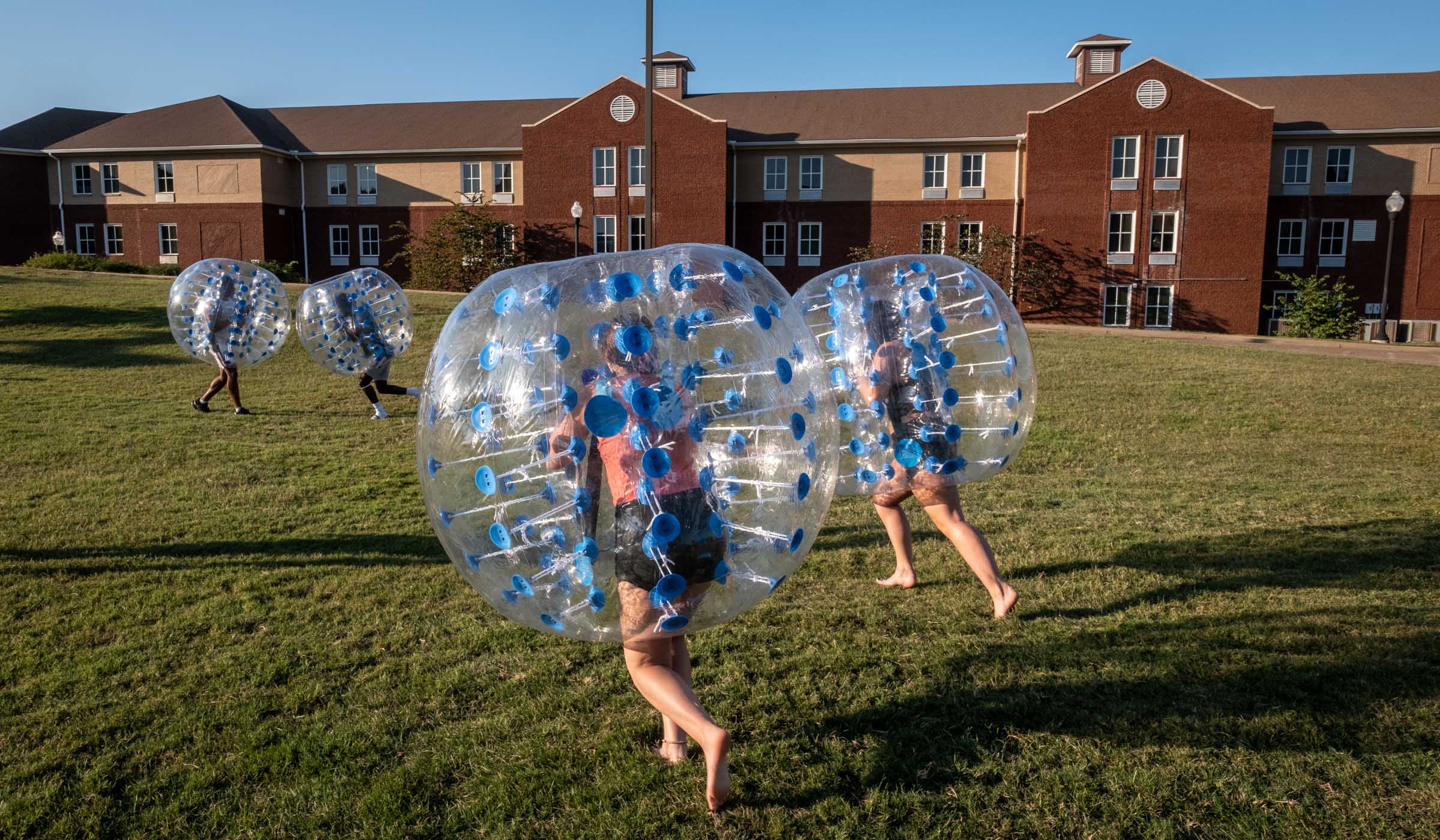 Students encased in inflatable cushione balls run towards eachother on a field in the setting sun, wih Ruby Hall in the background.