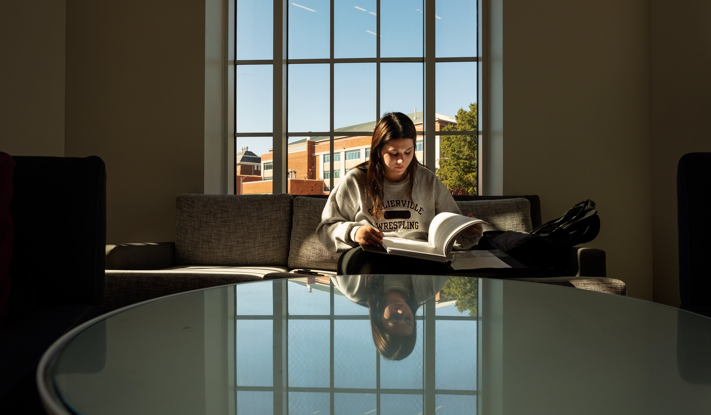 A student reads while sitting on a couch in the Rula atrium, with she and the window behind her reflected on a table in the foregound.