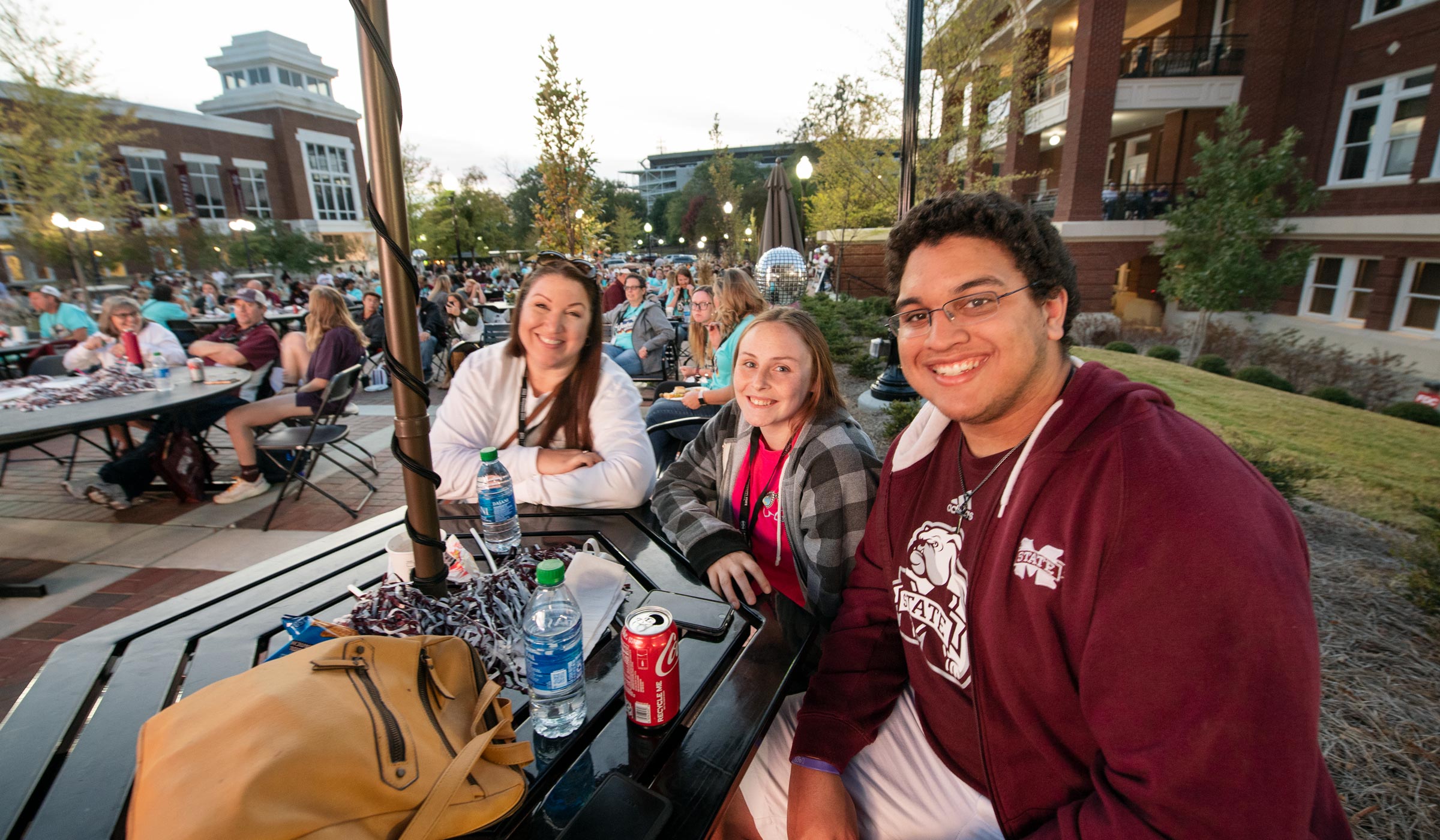 Three family members smile at the camera while sitting at an outdoor table on YMCA Plaza, with views to hundreds of others Super Bulldog Families beyond.