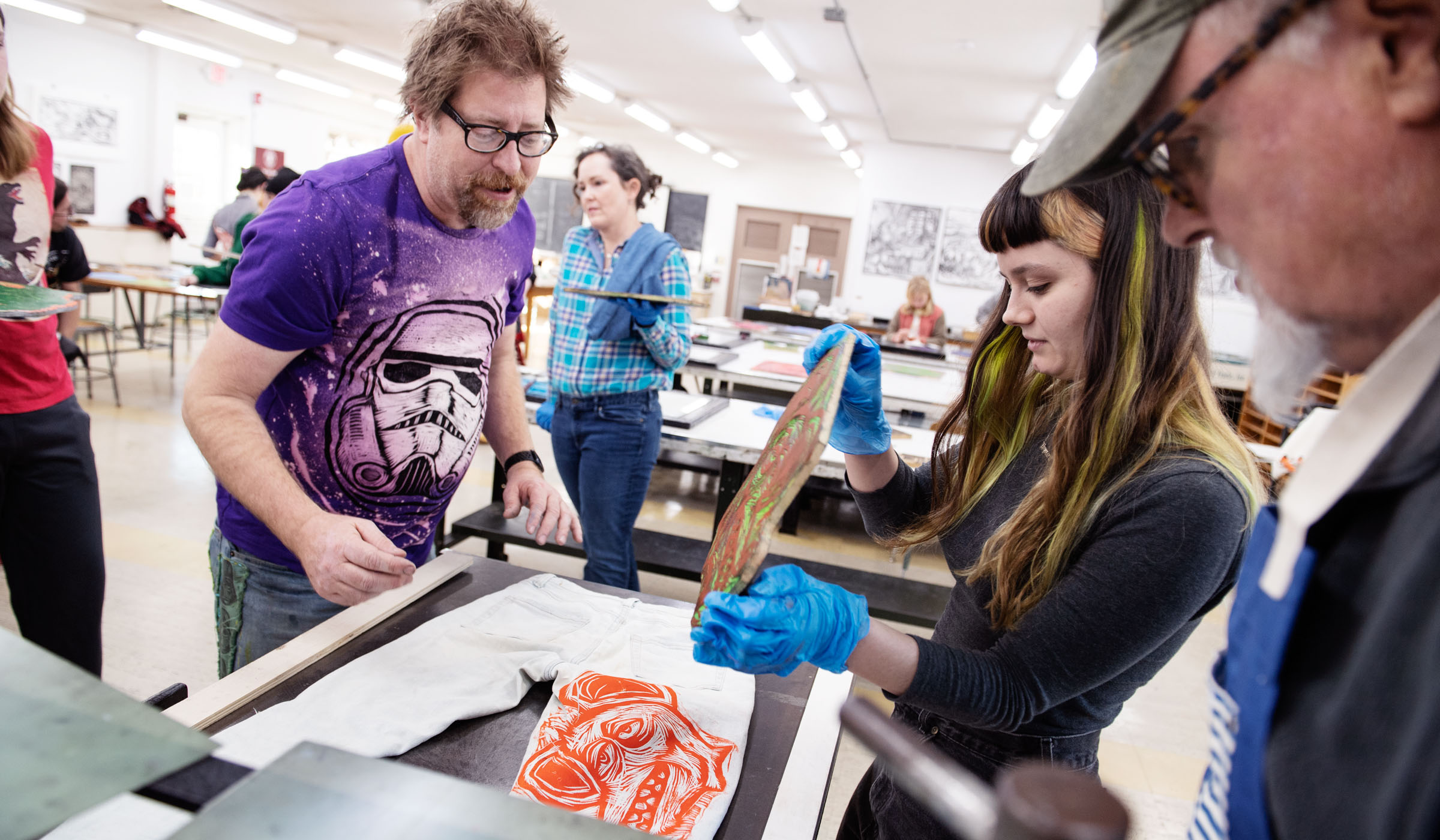 Visiting artist Sean Star Wars at left, wearing a t-shirt printed with one of his Storm Trooper helmet designs supervises Art student Juliet Buckholdt (right) pull an inked plate off, leaving an orange rat design on her pale jeans.