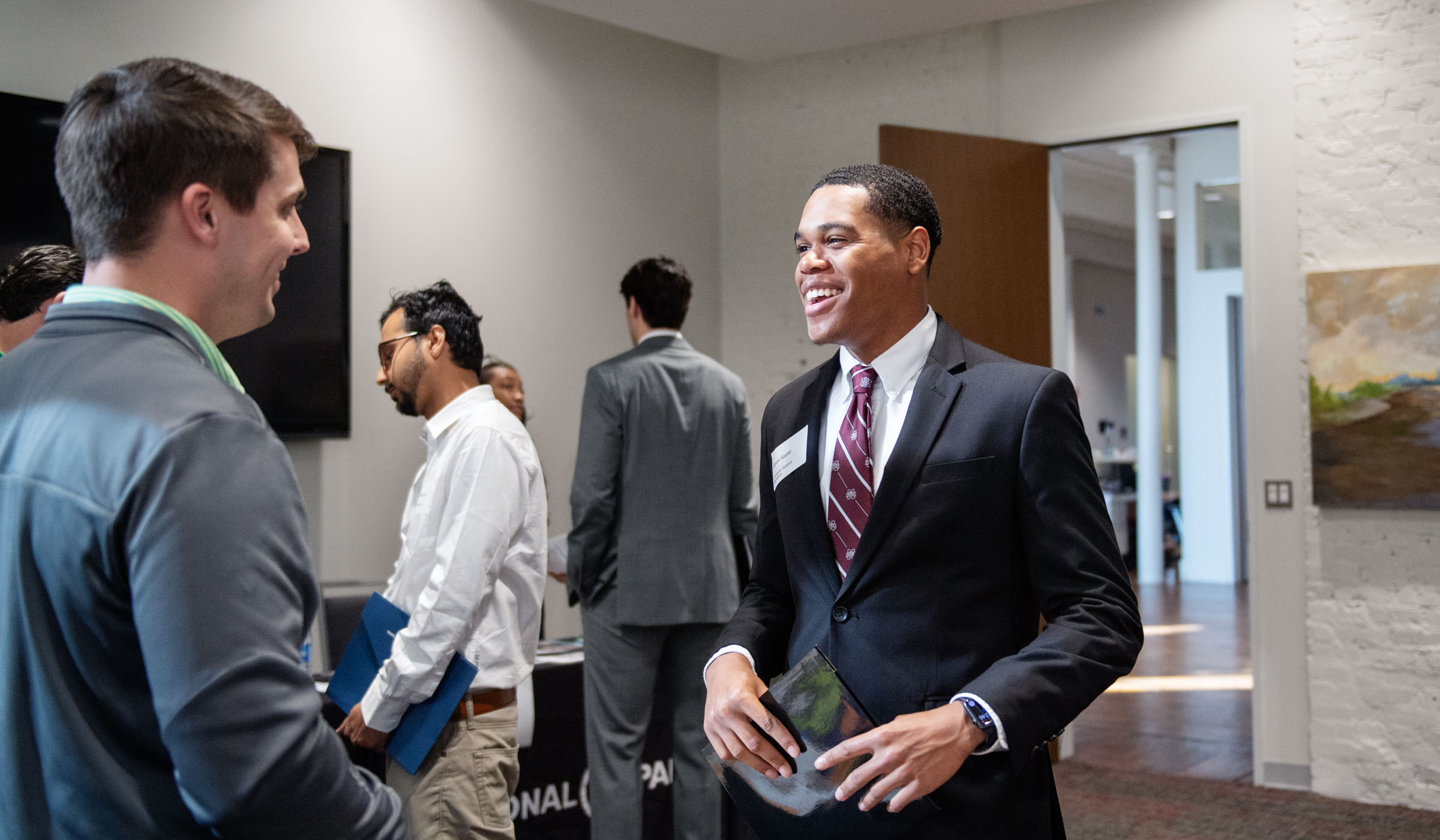 A student in a suit holding a portfolio folder at center smiles while talking to a prospective employer.