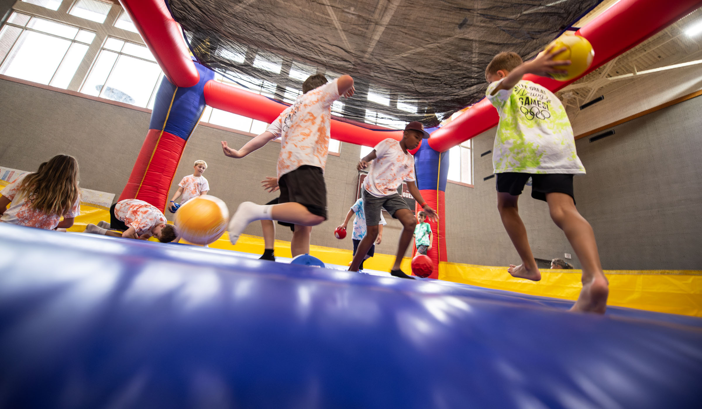 A bouncy castle with a blue inflatable floor and red supports is filled with children running and jumping with balls.