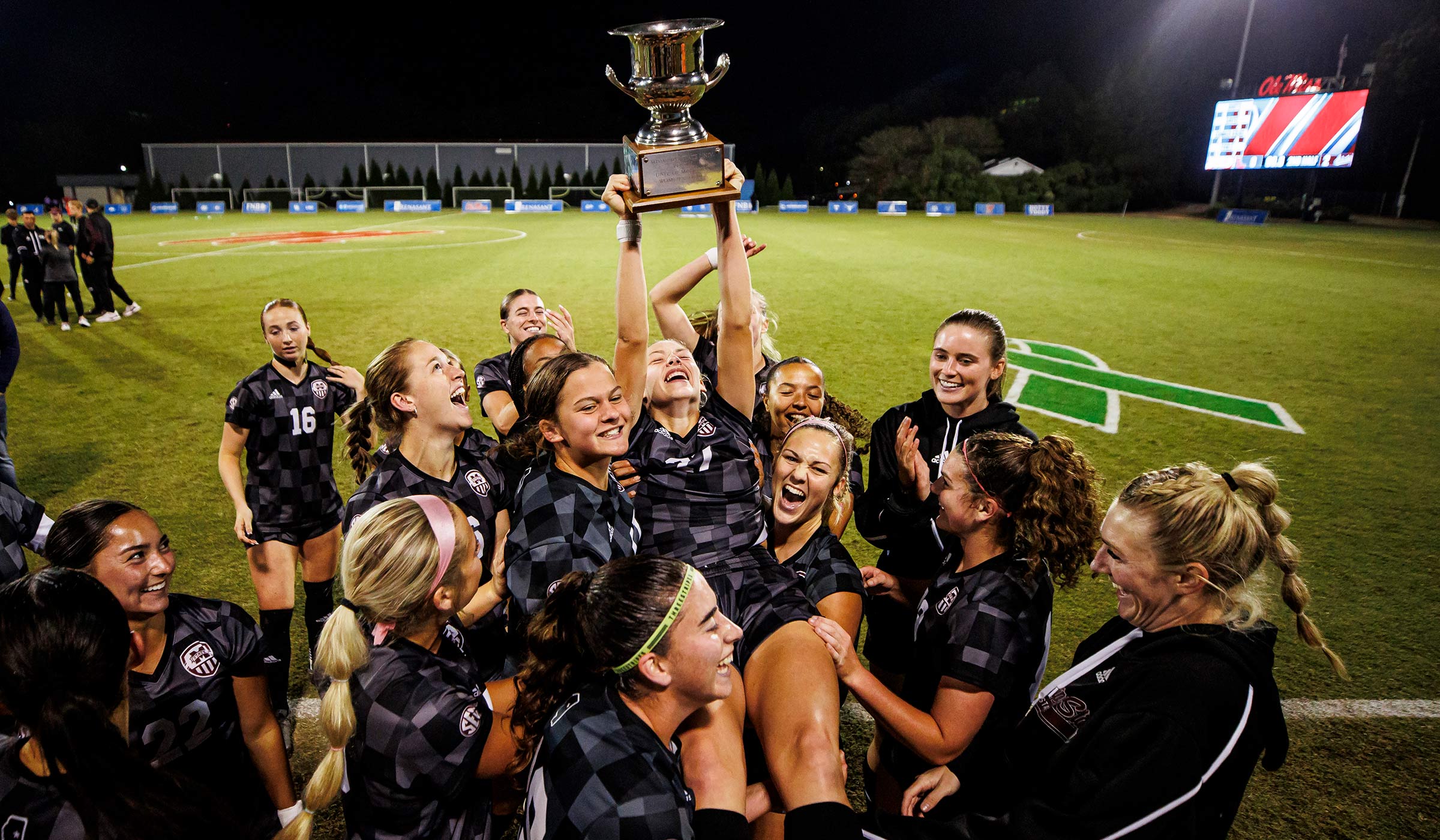 Soccer team in dark uniforms celebrating with player hoisted in air with trophy