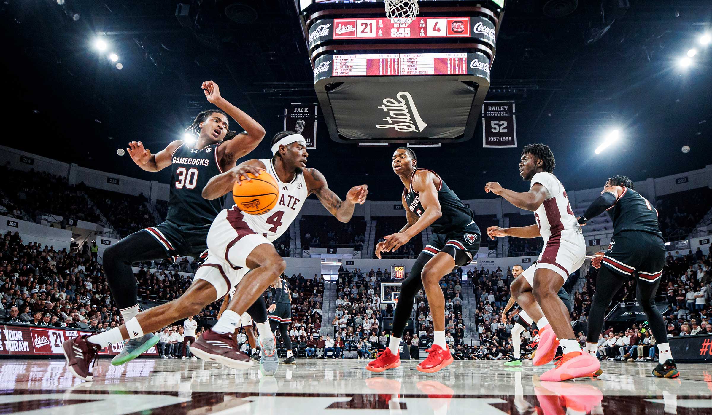 Basketball player in maroon and white dribbling near basket with defenders in black uniforms