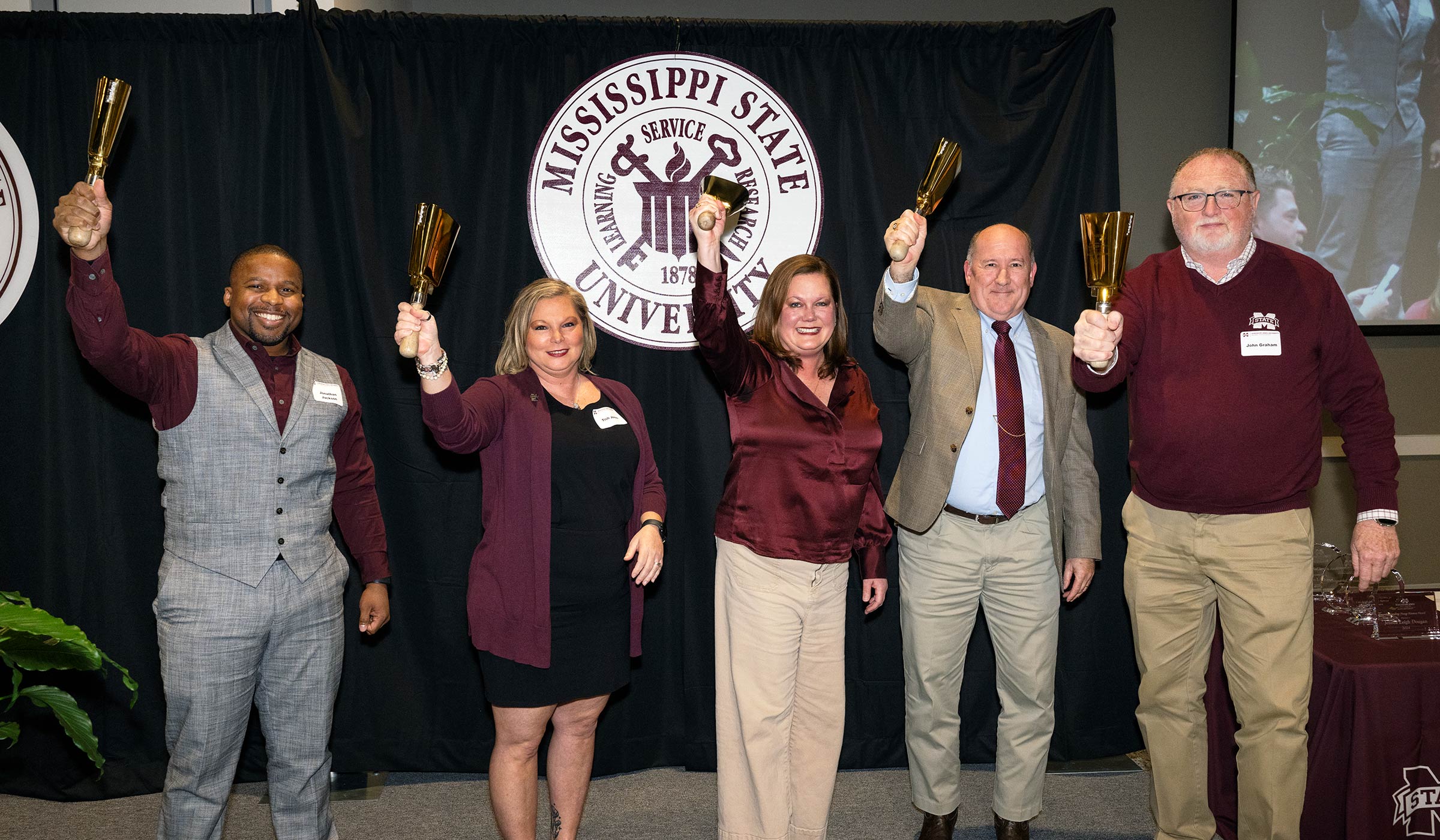 People wearing maroon ringing gold cowbells on stage in front of MSU seal