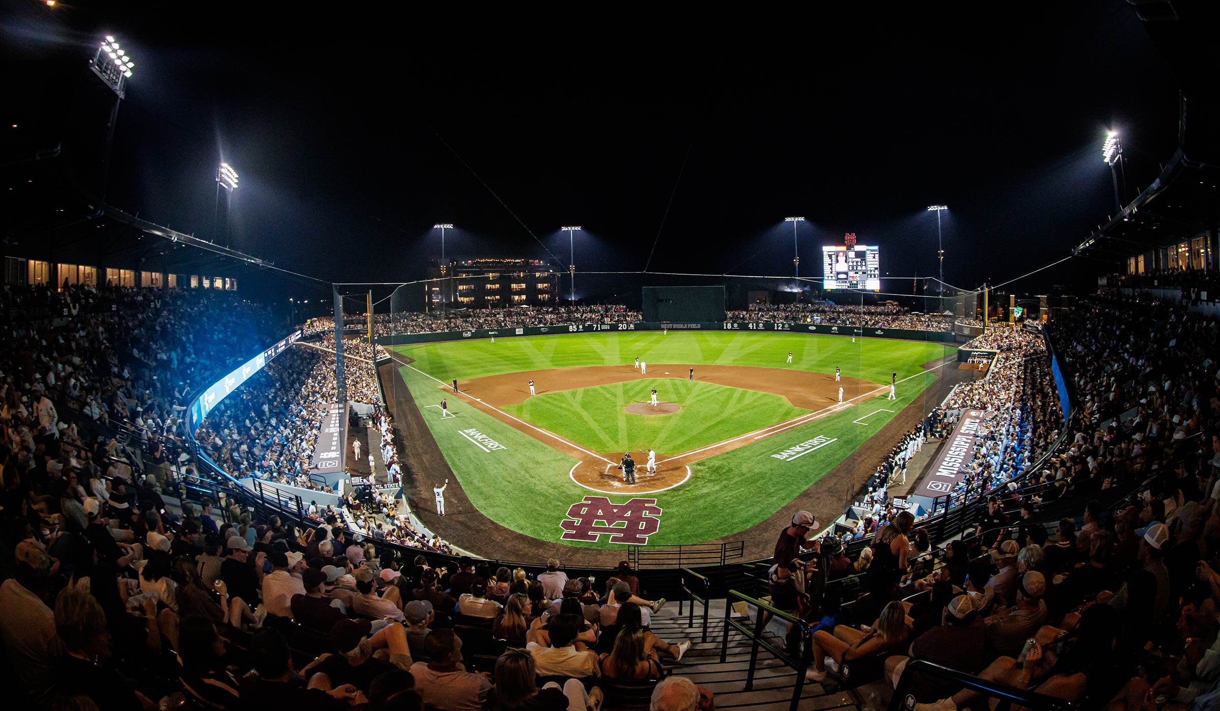 Baseball game at night under the lights with record crowd and bright green grass and dirt diamond and M over S logo in maroon