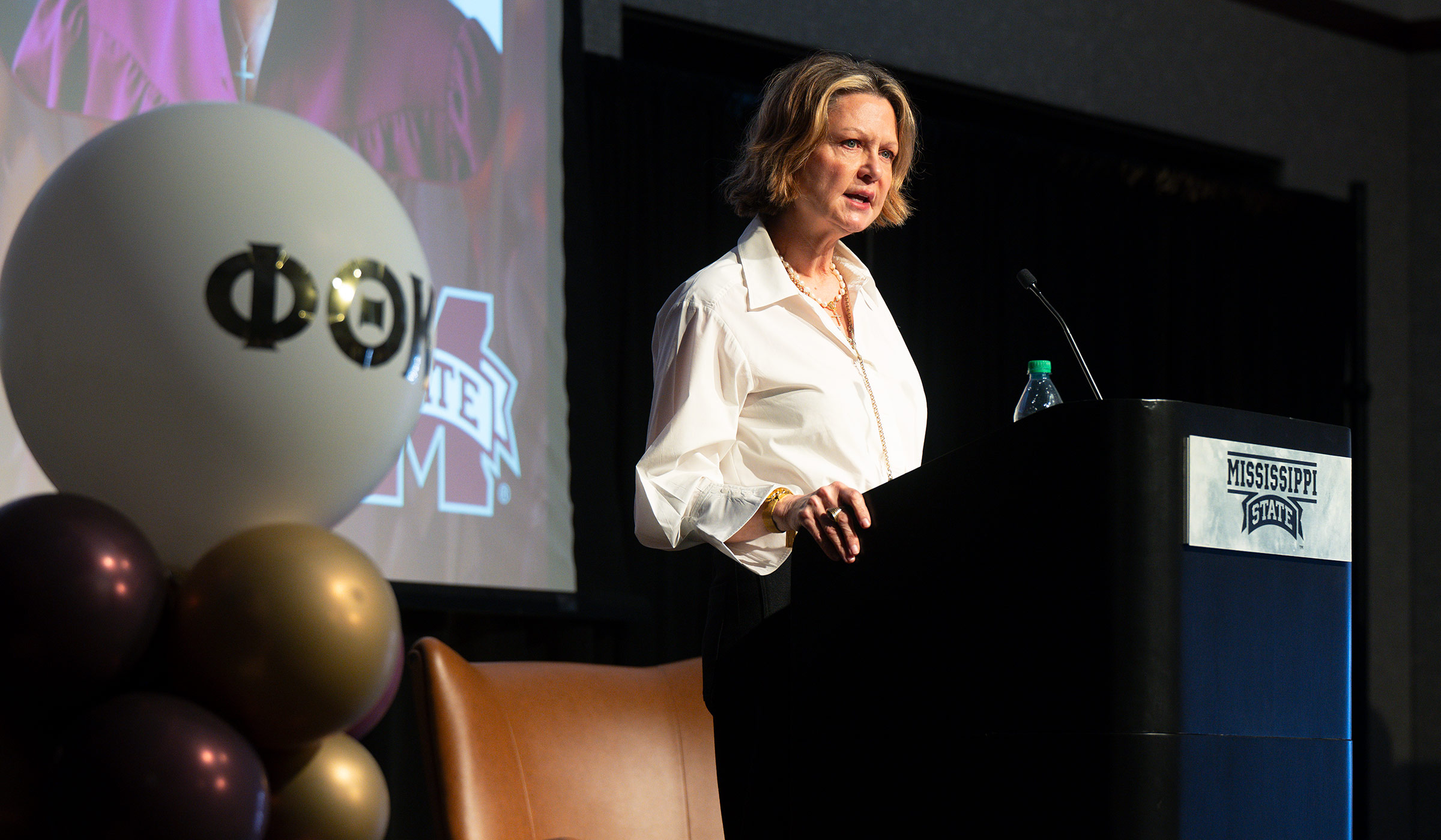 Woman in white shirt speaking at podium with balloon with Greek letters in foreground