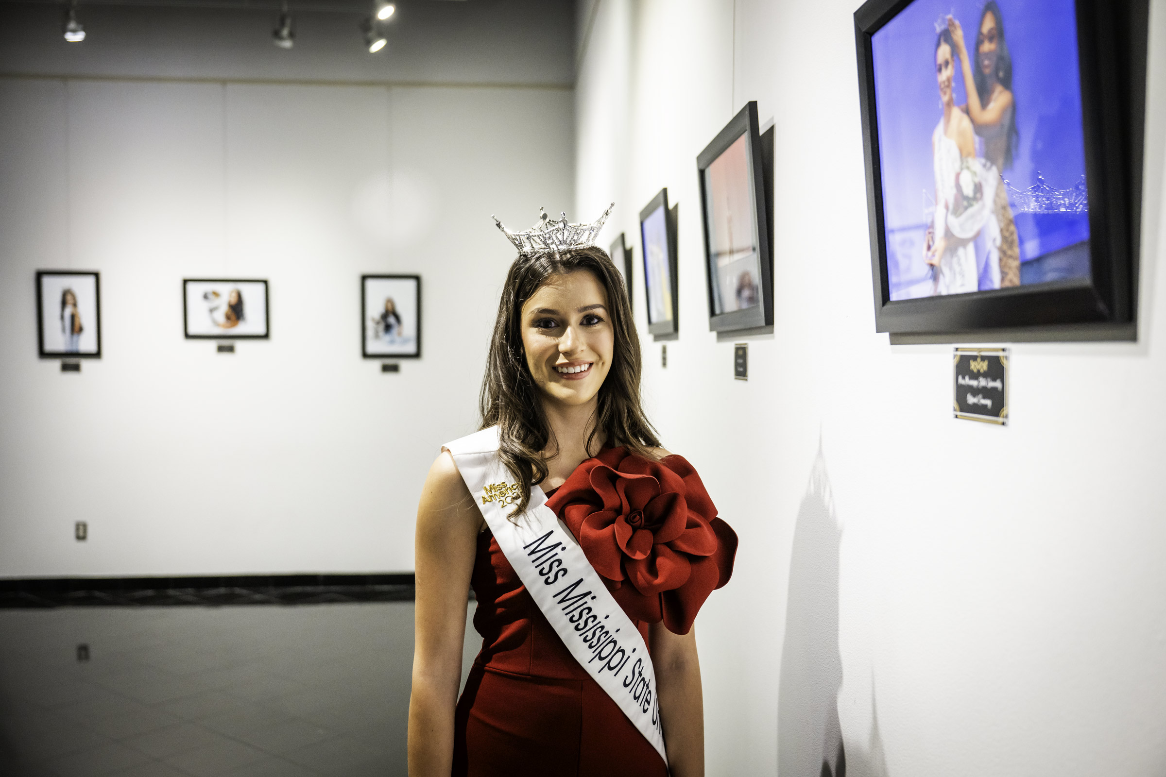 Miss Mississippi State University Karolina Heathcock smiles while wearing a deep red gown, crown, and official Miss MSU sash in an art gallery setting among multiple framed photos of her time onstage at the 2026 Miss MSU pageant. 