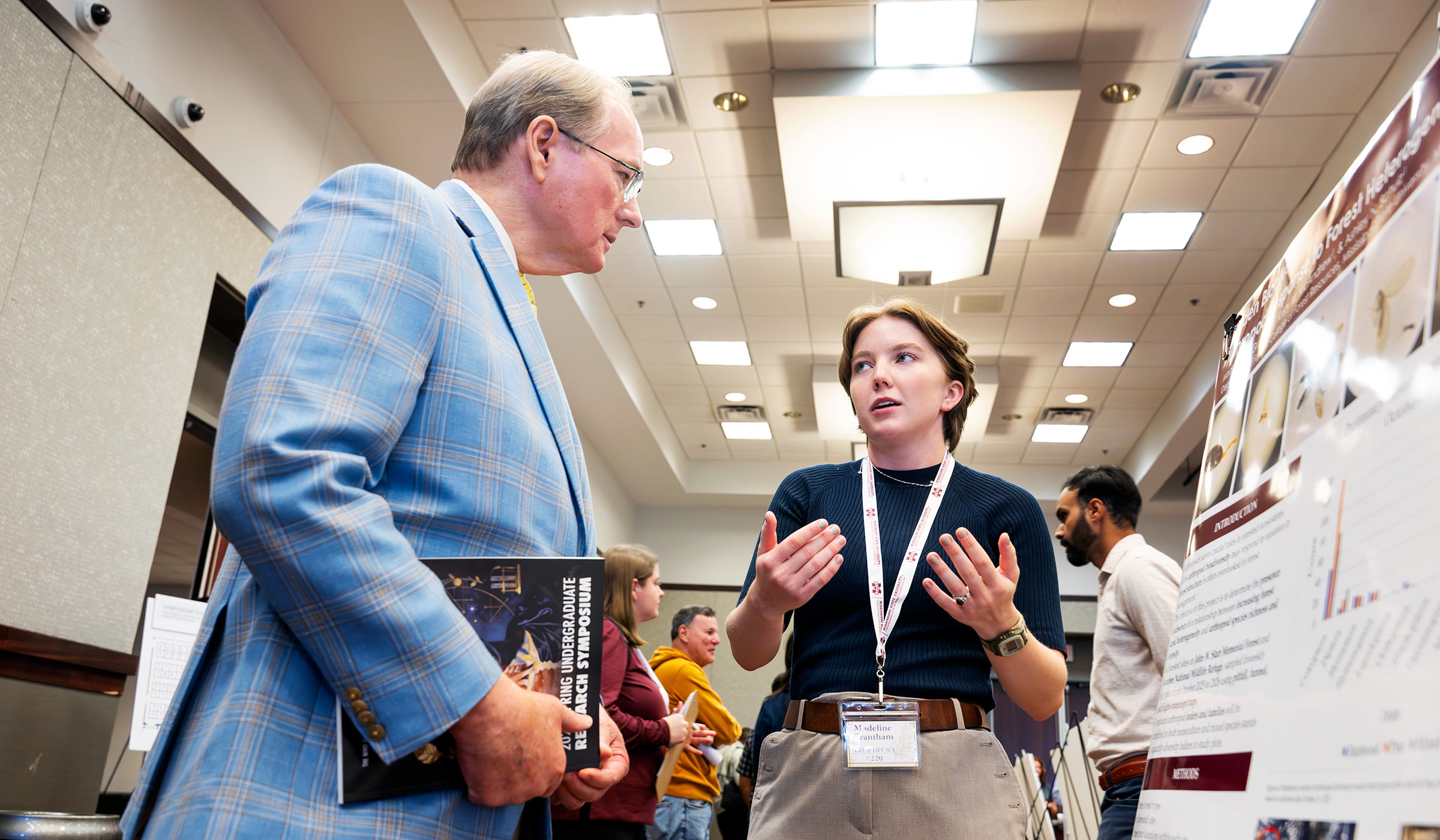 Man in blue blazer holding book speaking to female student in navy top and khakis discussing research presentation on poster board.