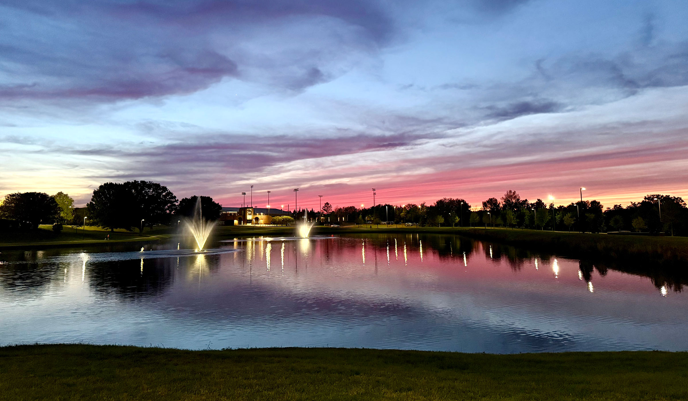 A purple and pink sunset over Chadwick Lake on campus is reflected in the water. The fountains in the lake are lit, as well as the softball and tennis complex in the background.