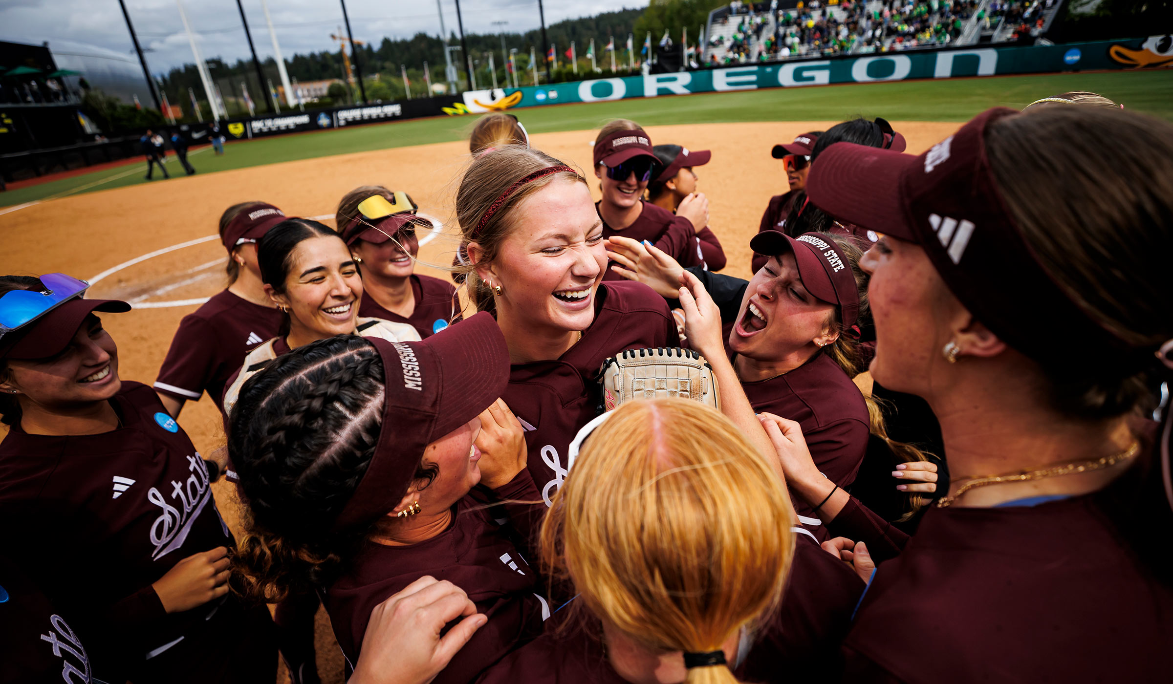 Women in maroon softball uniforms with white scripted state on front huddled together laughing and celebrating on a softball field with Oregon Ducks on the back wall.