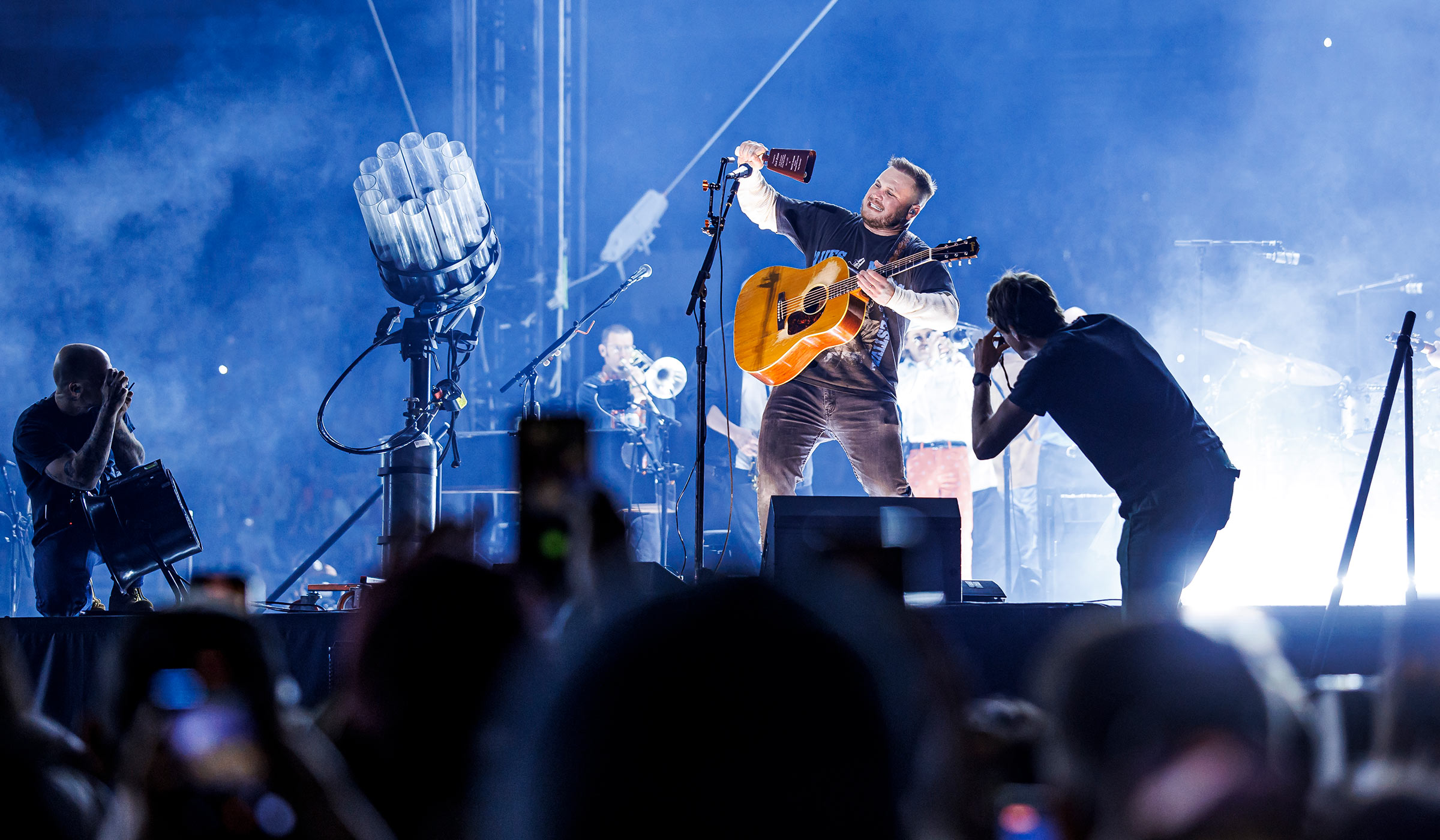 Two photographers in silhouette flanking a man on stage with a guitar and he is ringing a cowbell while people record on their phones.