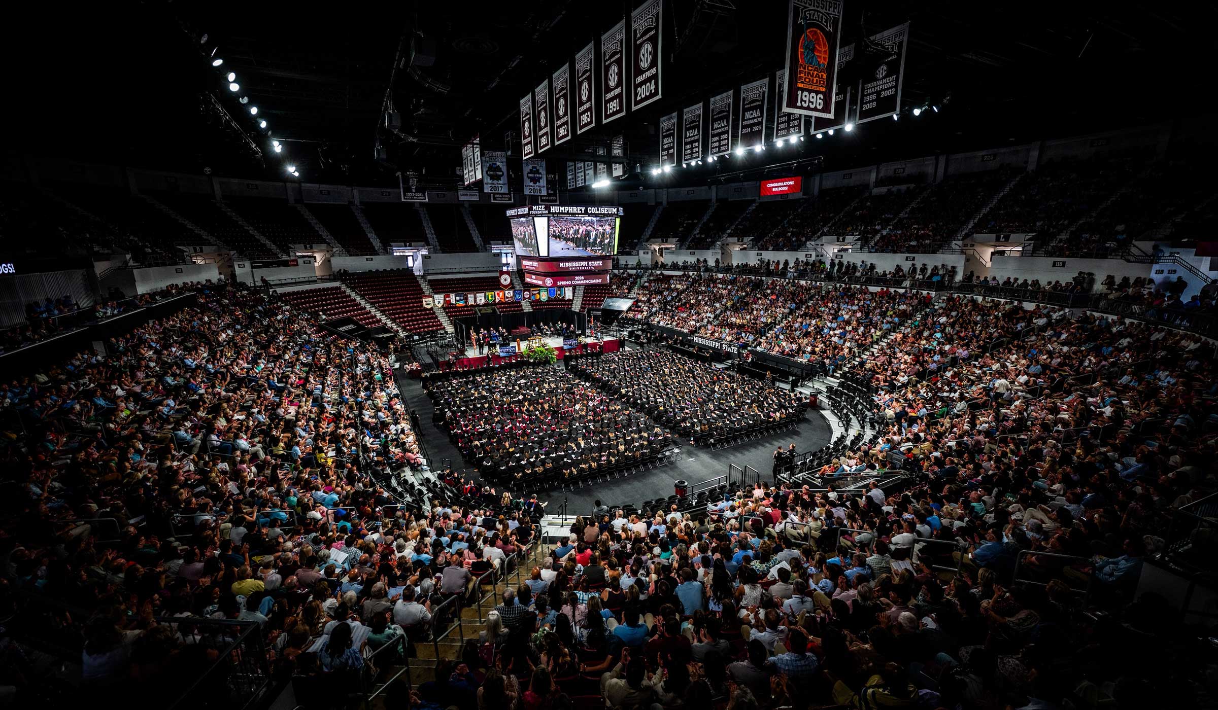 People sit inside of a basketball coliseum with maroon and white banners watching a decorated stage with colorful flags and people wearing black graduation regalia.