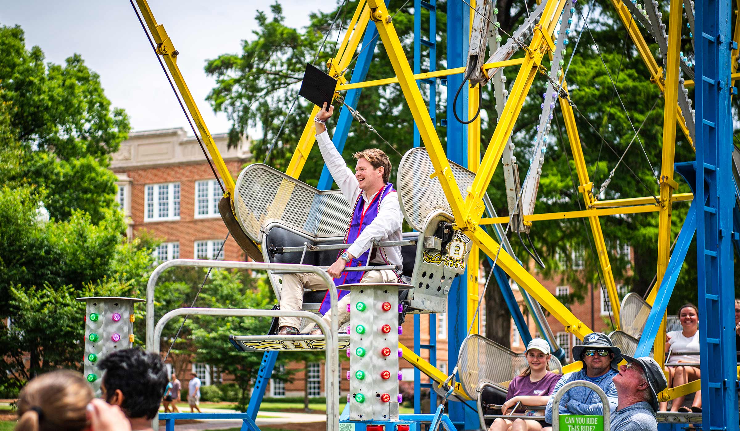 A student rides a Ferris wheel at a carnival on campus and waves his graduation cap in the air.