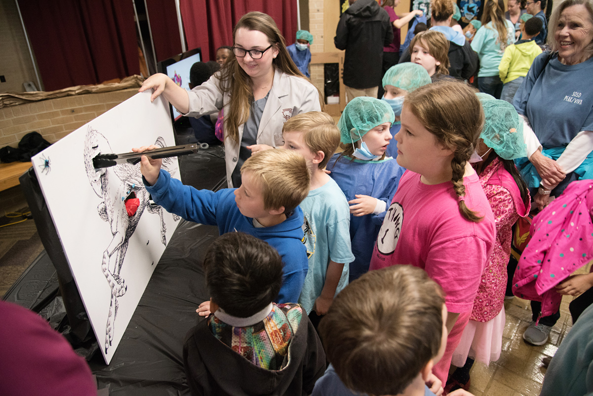 Child using giant tongs to perform pretend surgery on mystical animal painted on board.