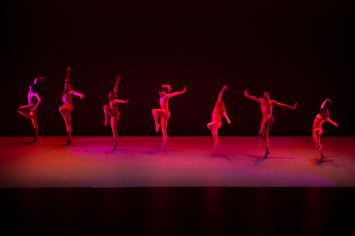 A row of seven dancers stretch across the stage, mid-action, lit with red and purple lights.