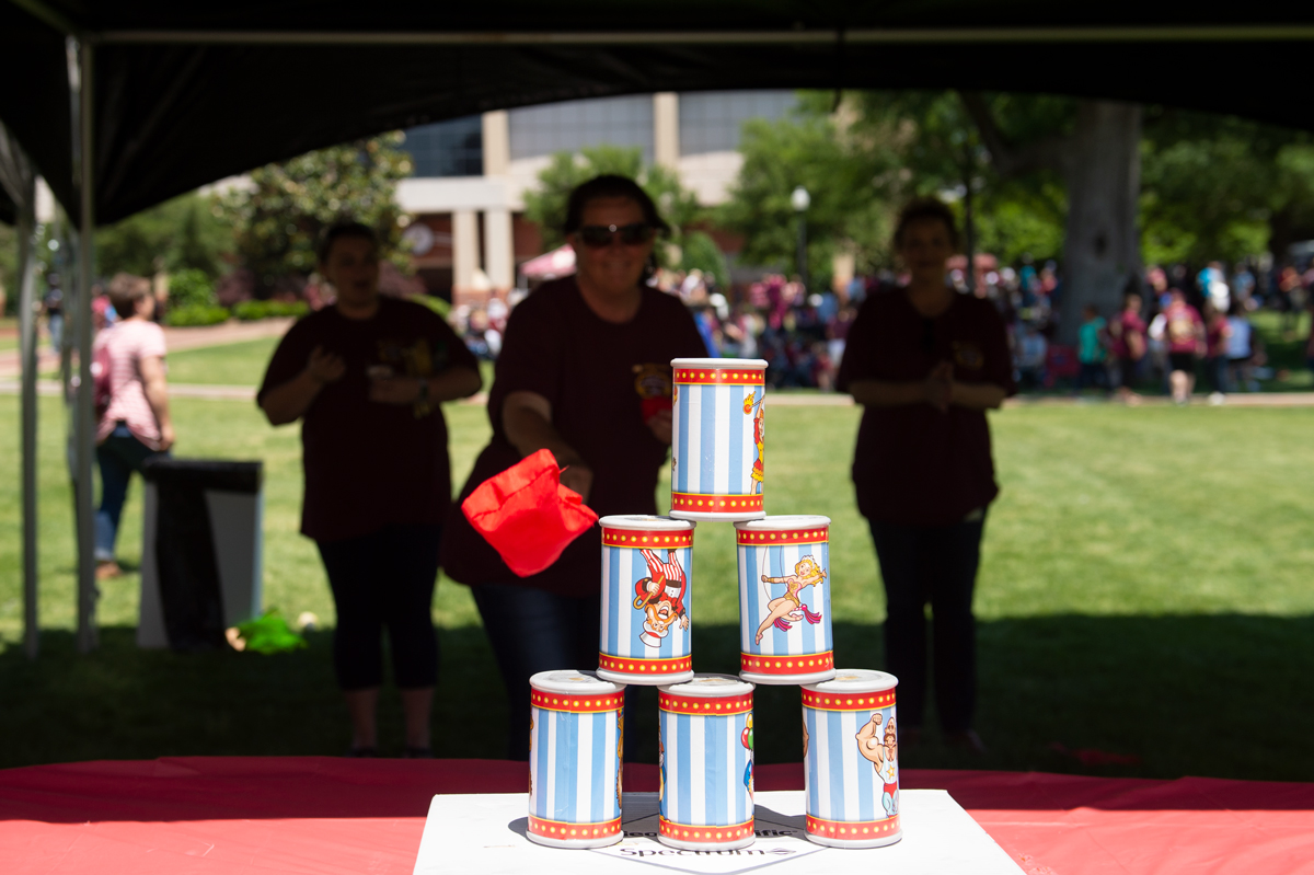 Three MSU staff members are sillhoutted under a tent while they take turns tossing a beanbag at a circus themed pile of cans.