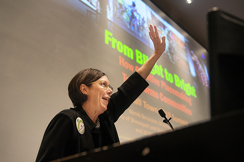 Wendy Benscoter gives a keynote address while standing at a podium in front of a large screen in Harrison Auditorium at Giles Hall.