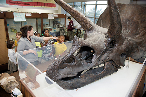 A scientific cast of a Triceratops skull at MSU’s Dunn-Seiler Museum in the foreground with children looking around the museum