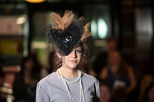 A student wearing a gray dress, white pearl necklace and black and gold lace hat with a bumblebee hanging down her left cheek stares at the camera.