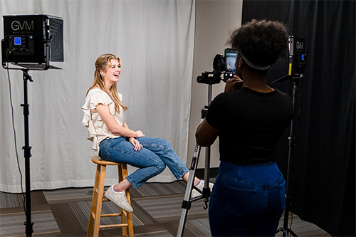 A student smiles for the camera and another student takes her picture in the new photography studio in Mitchell Memorial Library's MaxxSouth Digital Media Center.