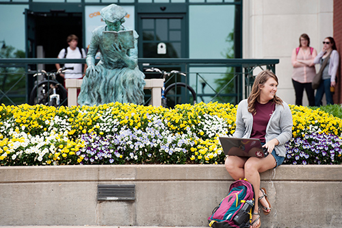 A student sits outside of Mitchell Memorial Library with her laptop, with flowers blooming and the "Scholar" statue in the background