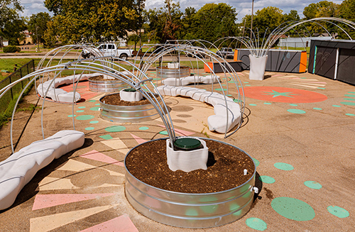 Raised garden beds and metal arbor structures are pictured in a fenced school area.