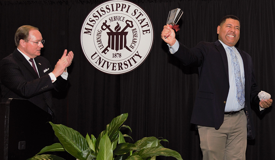 A man applauds as another rings a cowbell with the MSU seal in the background.
