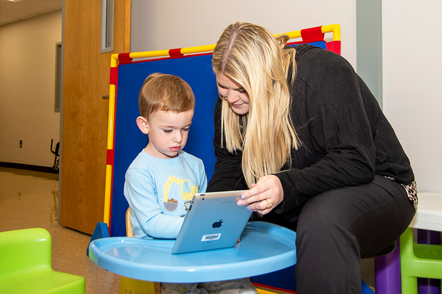 A young woman sitting on the right helps a young boy as they look at an iPad screen together.