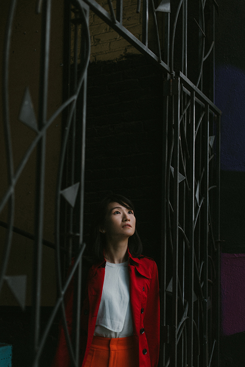 An Asian woman in a bright red coat stares up at the sky while standing near a large, black metal gate.
