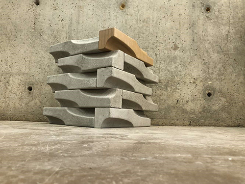 One tan and nine gray concrete masonry units stacked near a concrete wall