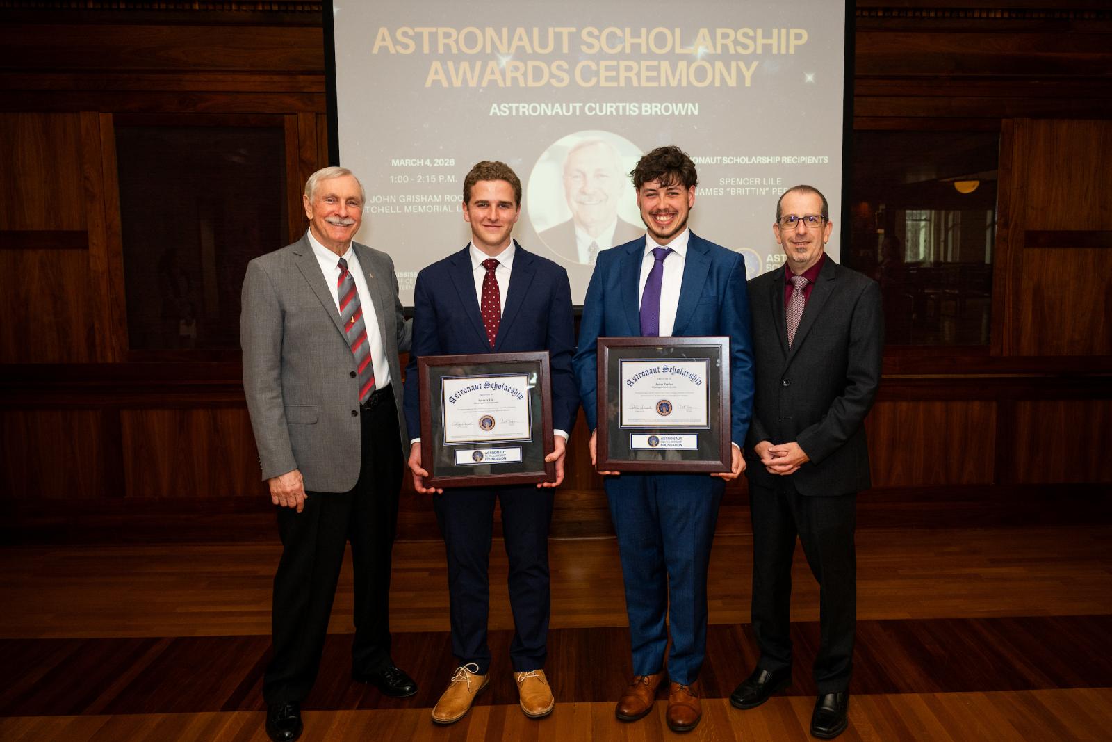four men, two with plaques in the center, stand in front of a backdrop