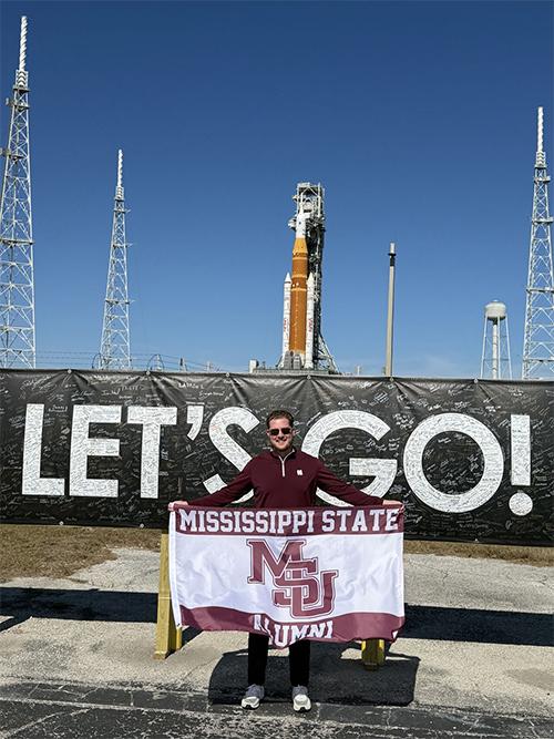 Parker Glass hold an MSU alumni flag in front of the NASA launch pad