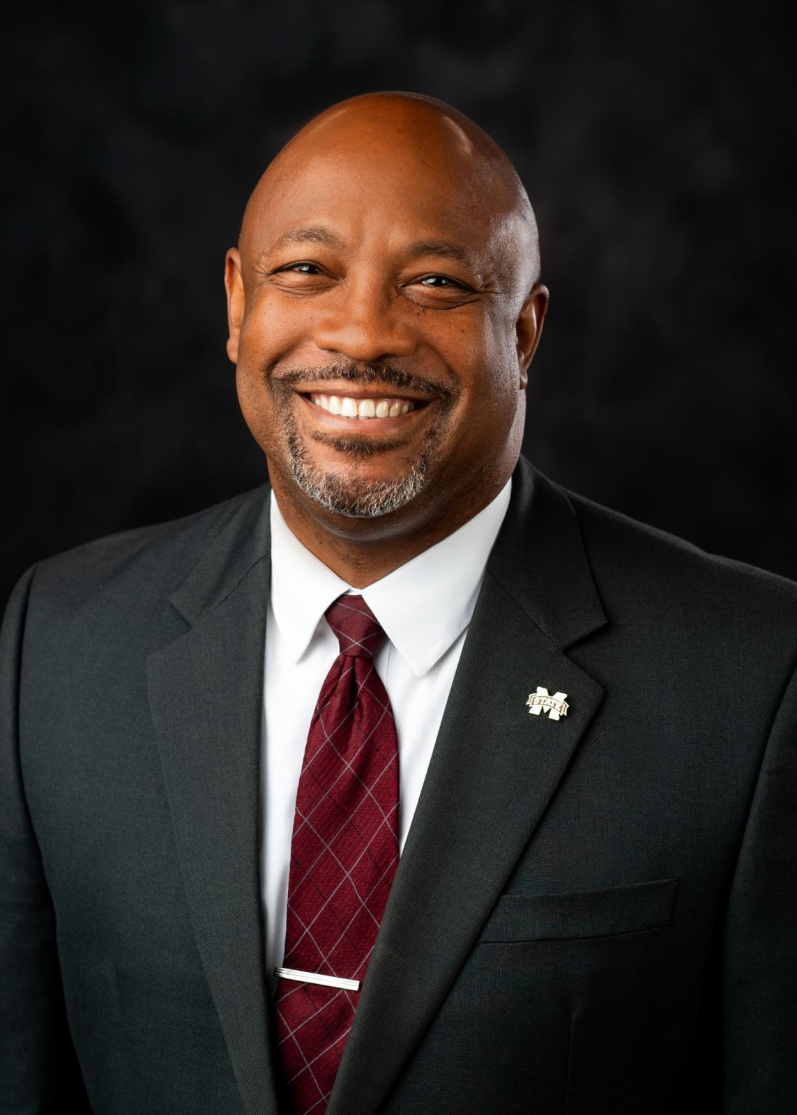 Headshot of Calvin Mosley in a black suit and maroon tie on dark background